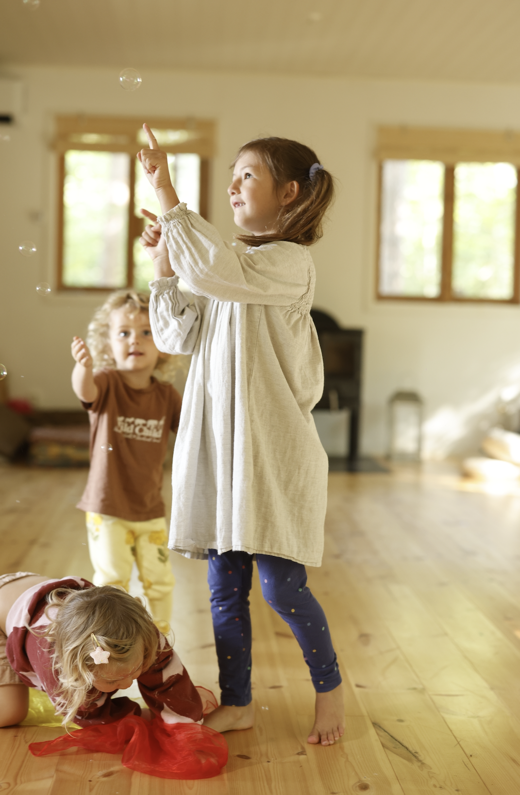 Children playing indoors, with one girl standing and pointing at bubbles, two girls on the floor playing with red fabric, and another girl in the background, in a well-lit room with wooden floors and large windows.