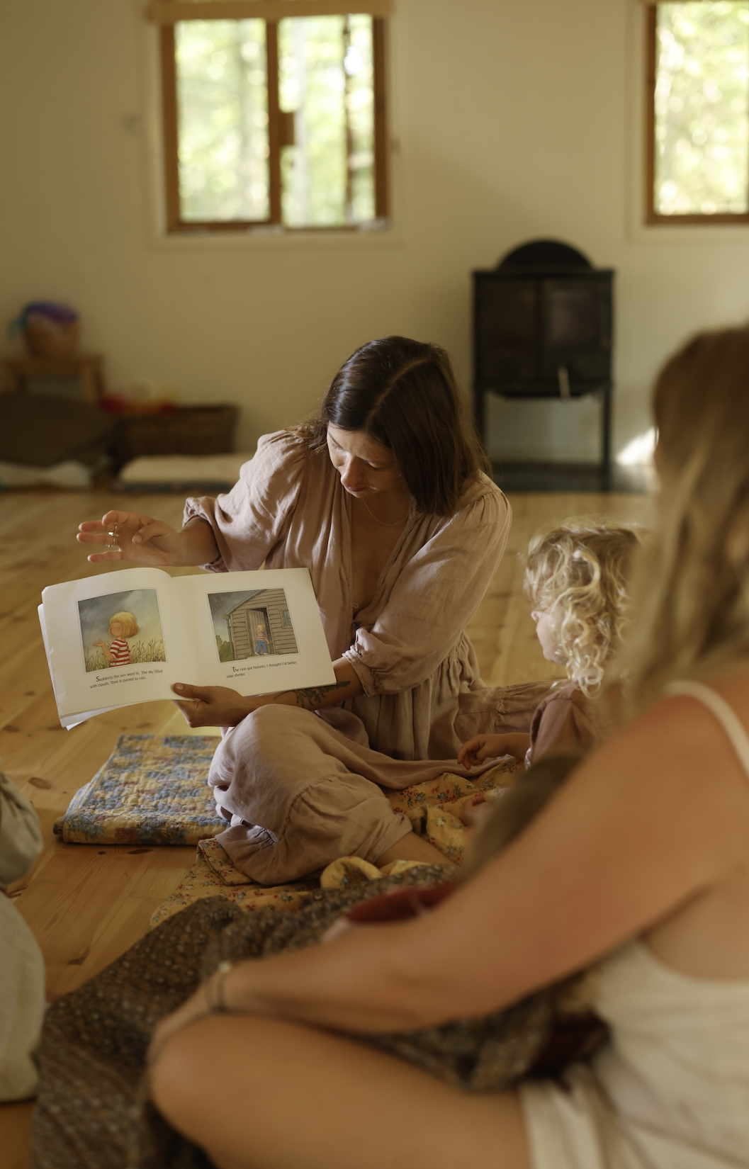 A woman reading a children's picture book to two girls in a cozy room with wooden floors and windows.