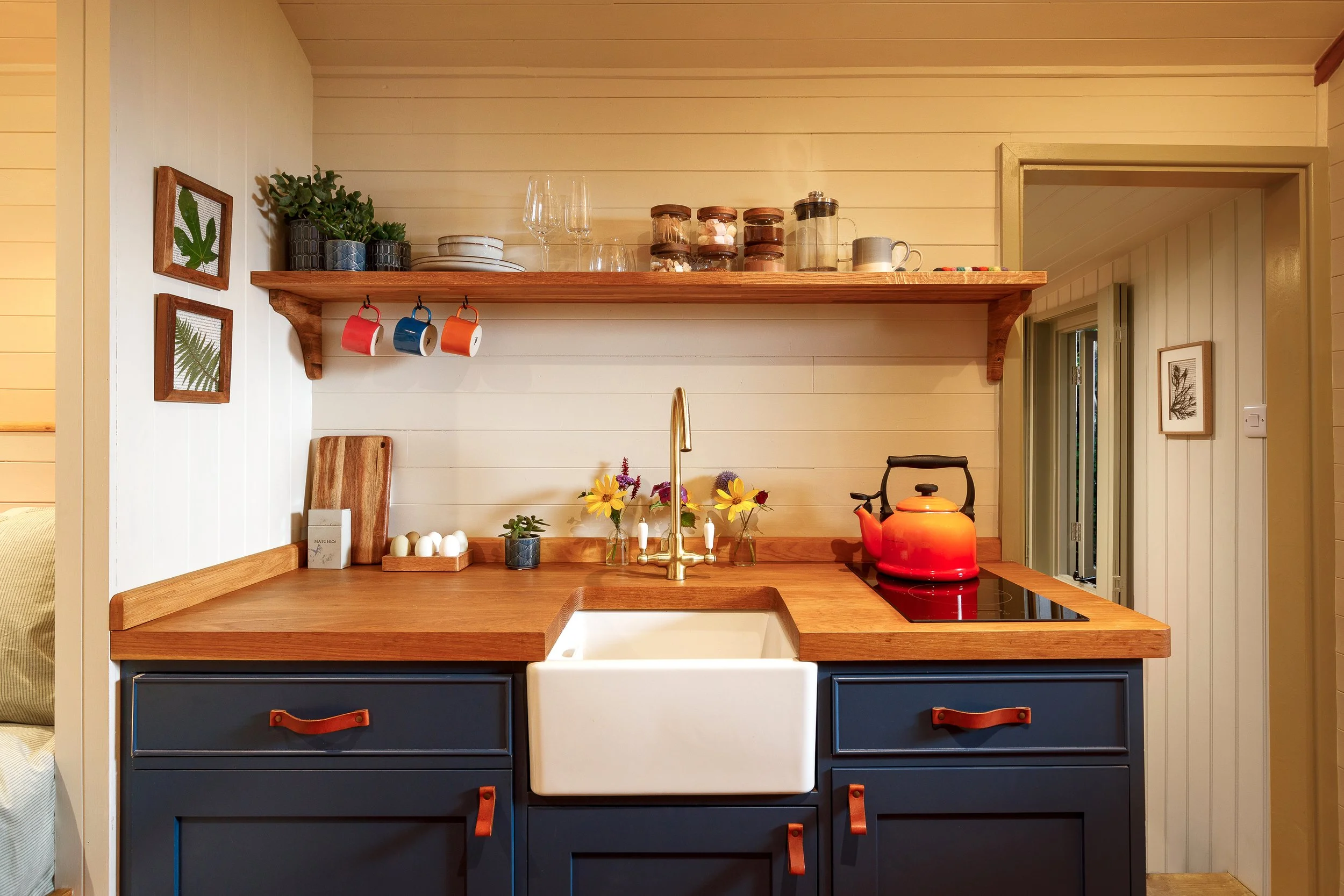 A cozy kitchen with a wooden countertop, navy blue cabinets with leather handles, a white farmhouse sink, a red kettle, and decorative plants and framed art on the walls.