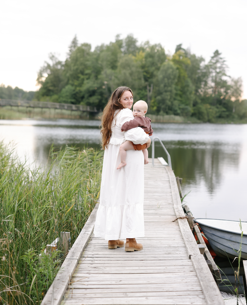 A woman holding a baby standing on a wooden dock beside a body of water with trees in the background.