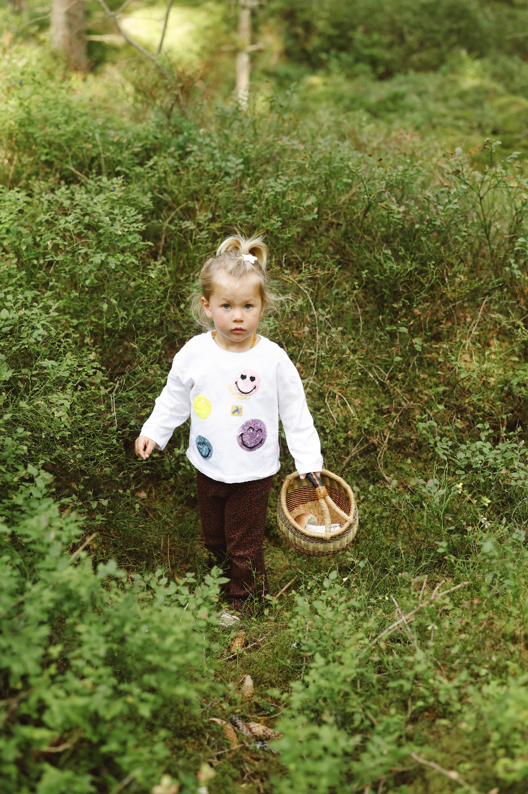 A young girl with blonde hair in a ponytail, wearing a white shirt with colorful smiley faces, dark leopard-print pants, and holding a wicker basket, walking through a dense green forest with bushes and trees.