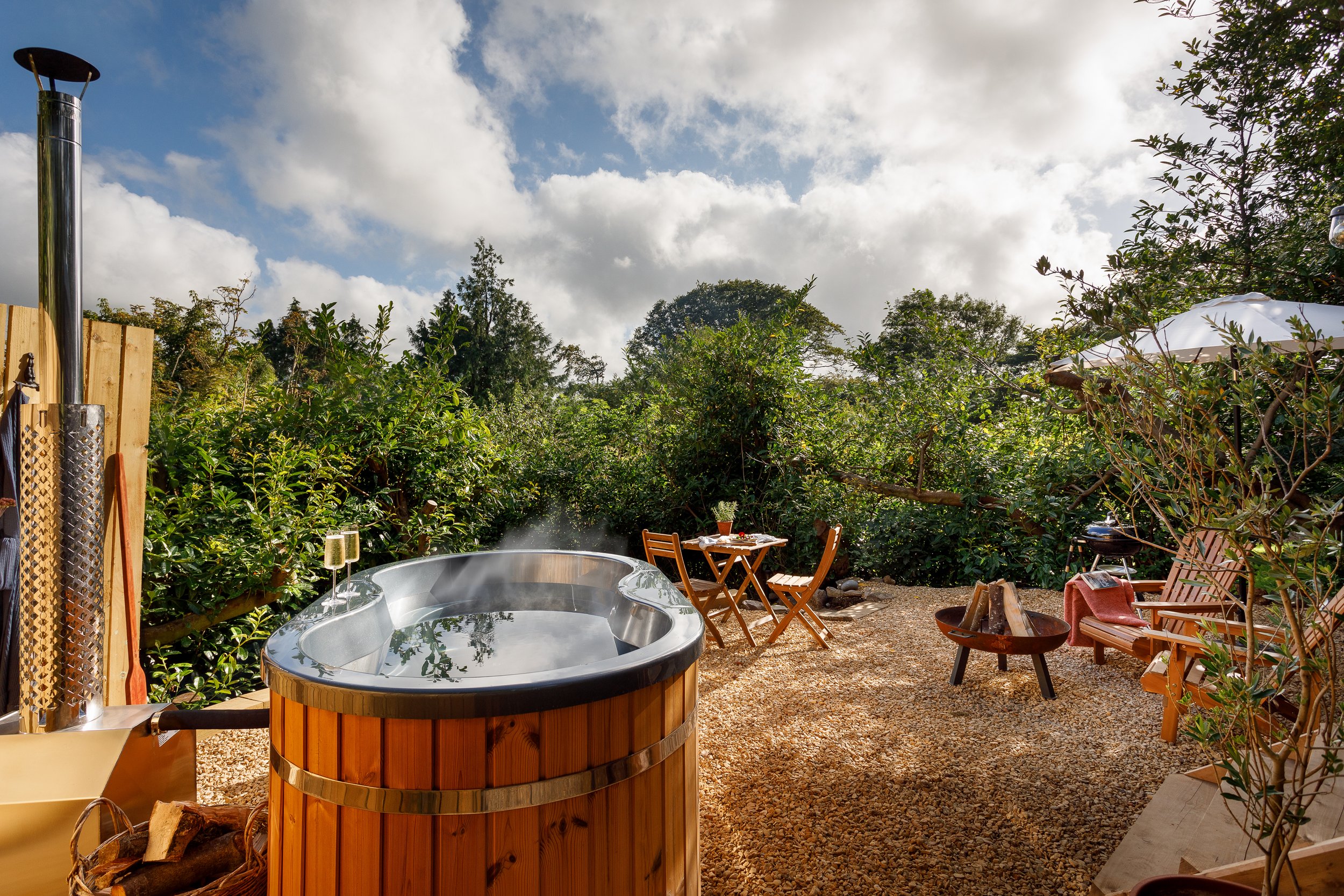 Outdoor backyard with hot tub, wooden deck, small table with two chairs, seating area with a fire pit, barbecue grill, and lush green bushes under a partly cloudy sky.