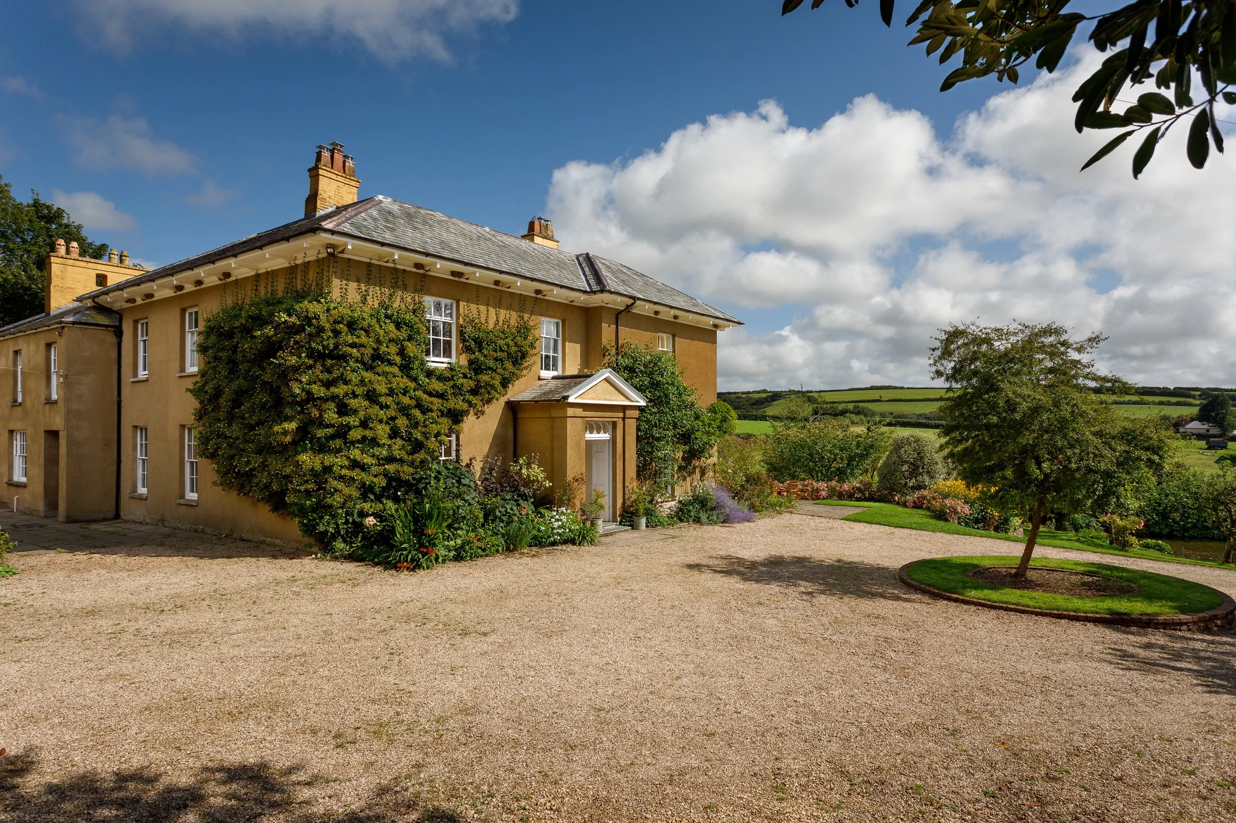 A yellow two-story house with a gray roof, surrounded by a gravel driveway, green trees, and a lush garden with flowering bushes in a rural setting under a partly cloudy sky.