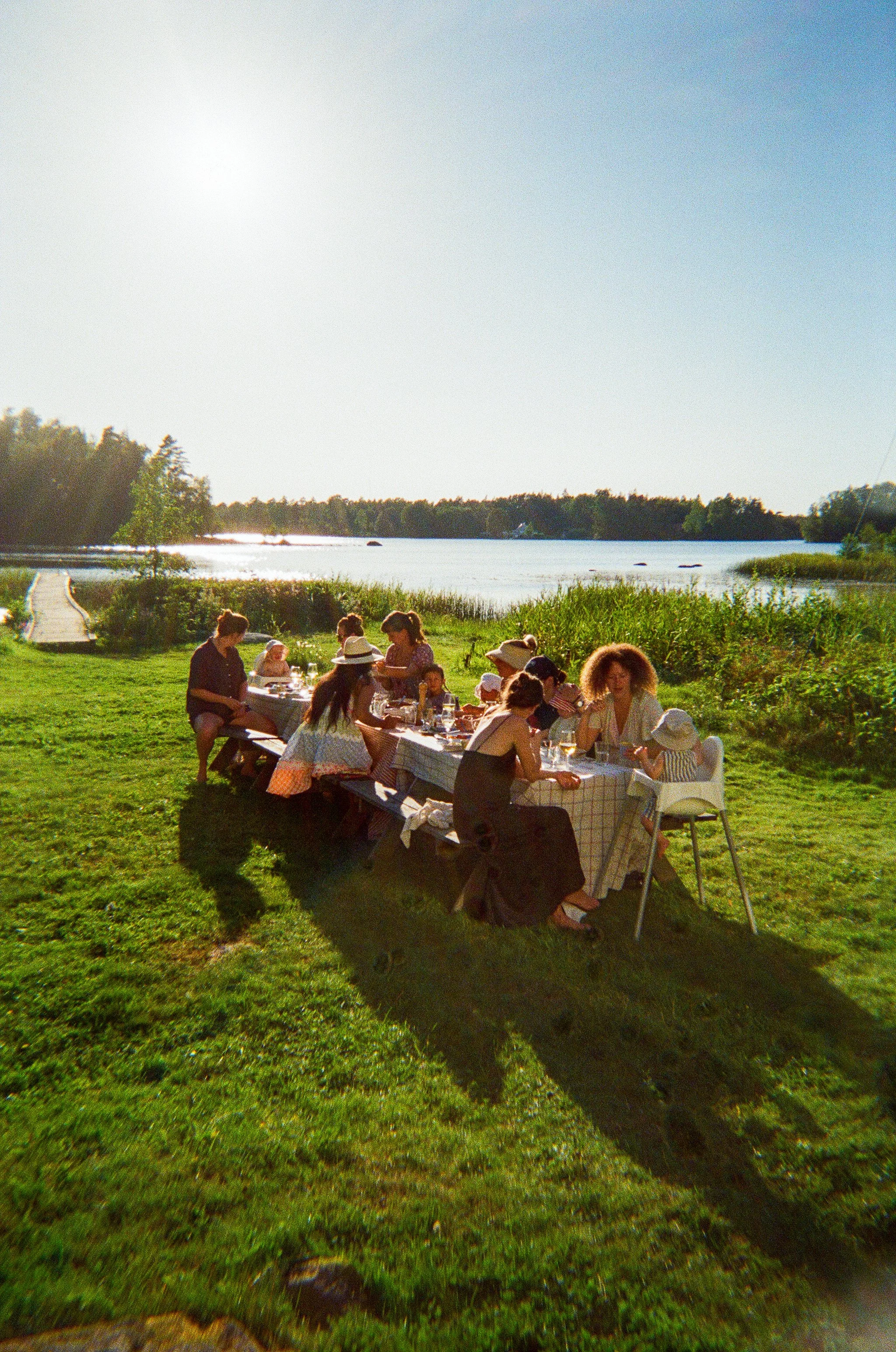A group of people, including children, having a outdoor meal at a long table on a grassy area near a lake with trees in the background on a sunny day.