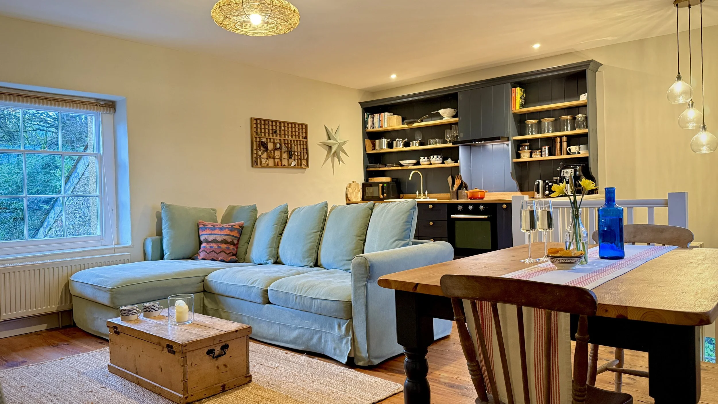 Living room with a large window, beige sectional sofa with colorful pillow, wooden coffee table with candles, open kitchen with black cabinets, open shelving with dishes, dining table with blue vase and glasses, and modern pendant lights.