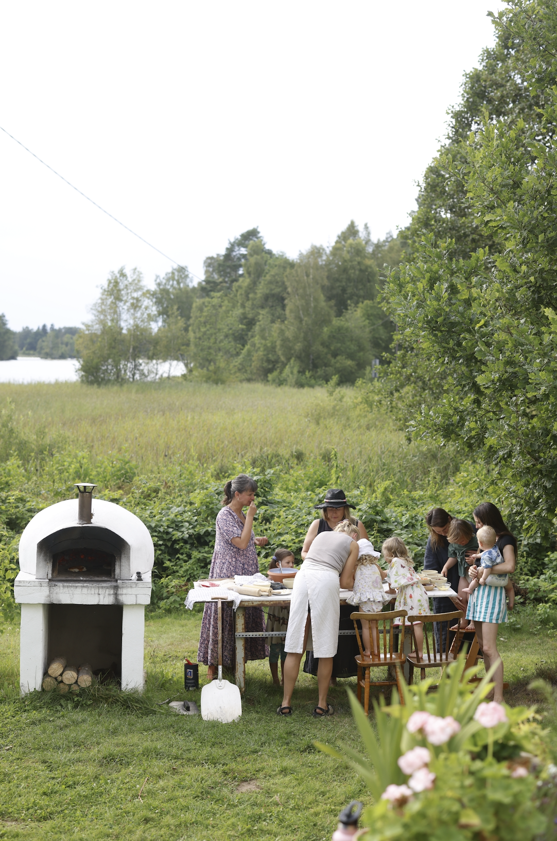 A group of women and children having a picnic or outdoor gathering in a grassy area near a pond or lake, with a white outdoor pizza oven and a green, tree-filled background.