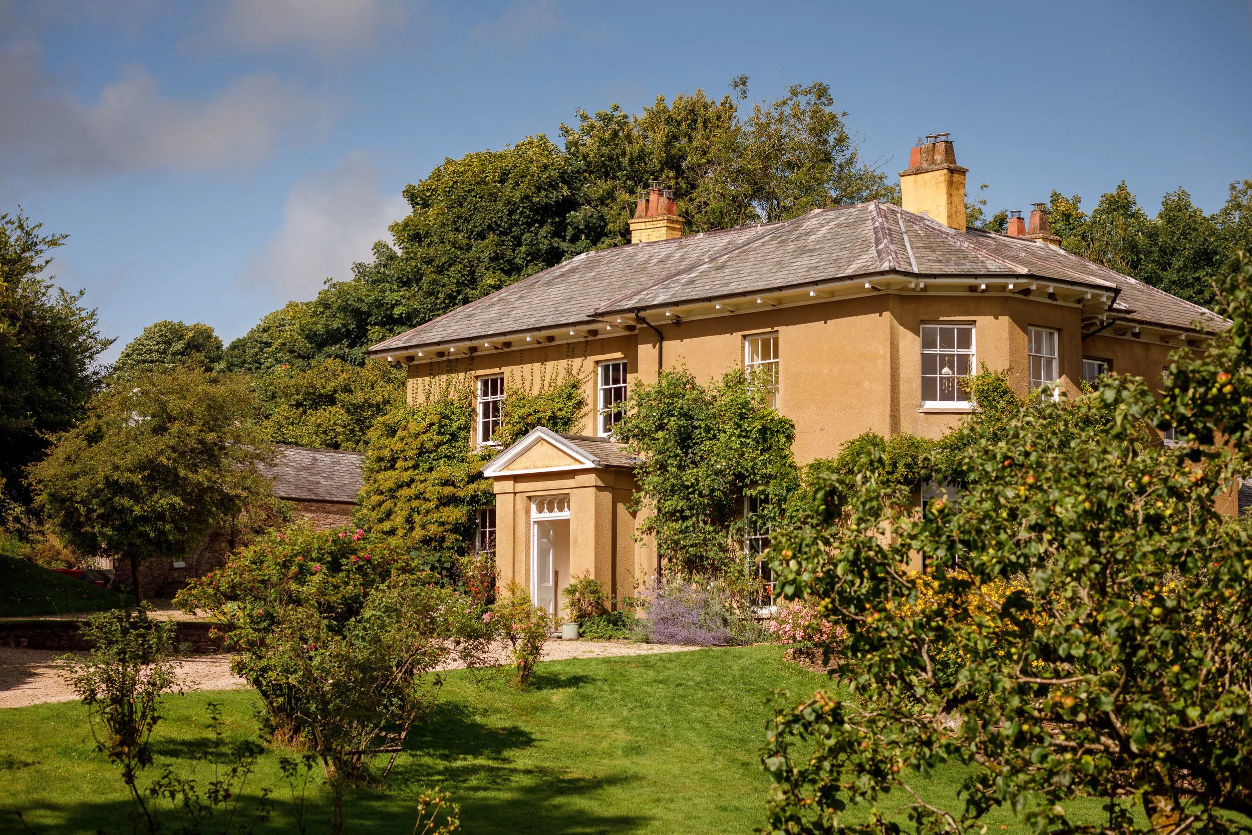 A two-story beige house surrounded by green trees and bushes, with a lawn in the front yard, under a partly cloudy sky.