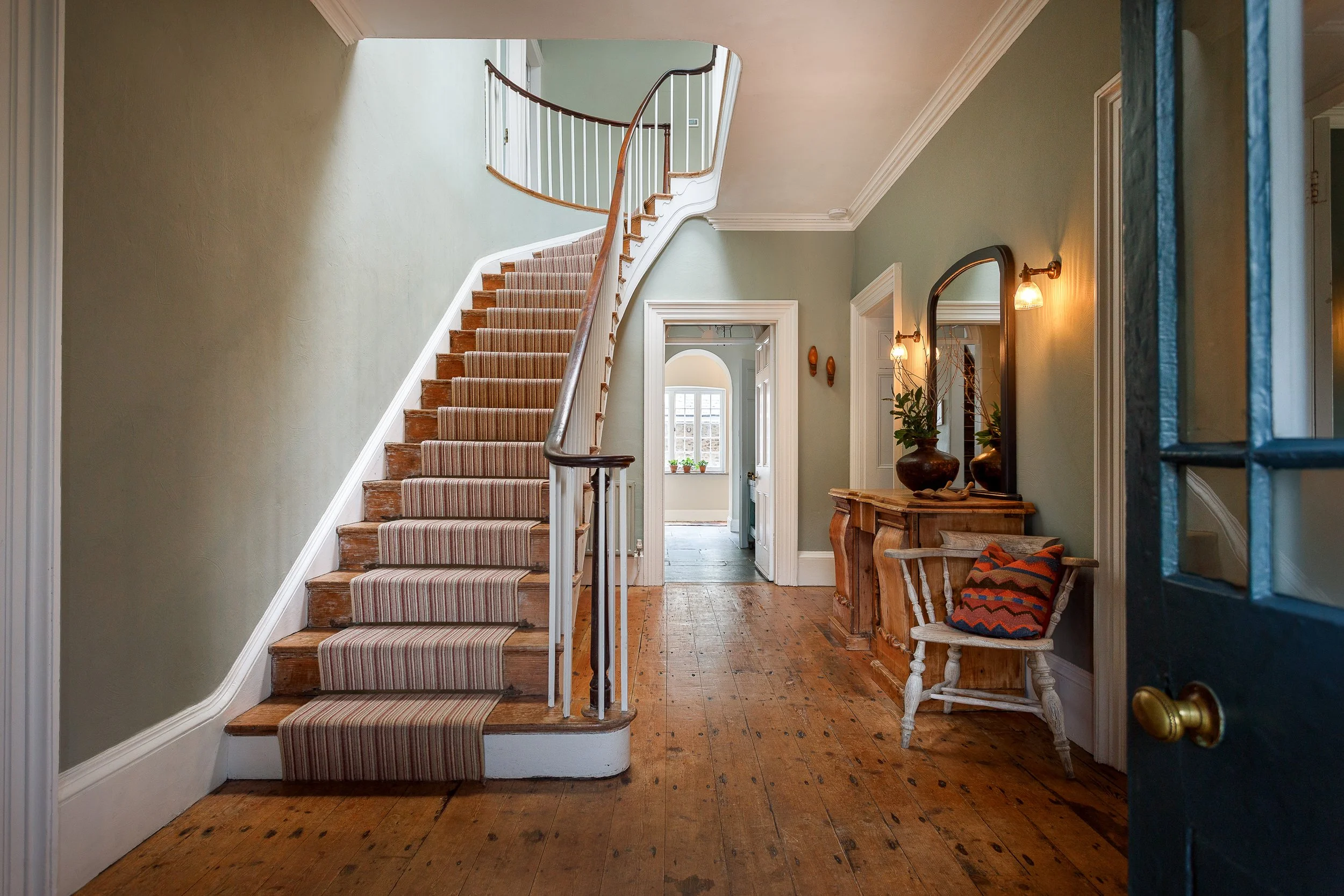 Interior view of a house entryway with wooden flooring, a staircase with striped carpet, a wooden console table with vases and a mirror, and a bench with a throw pillow, seen through an open door.