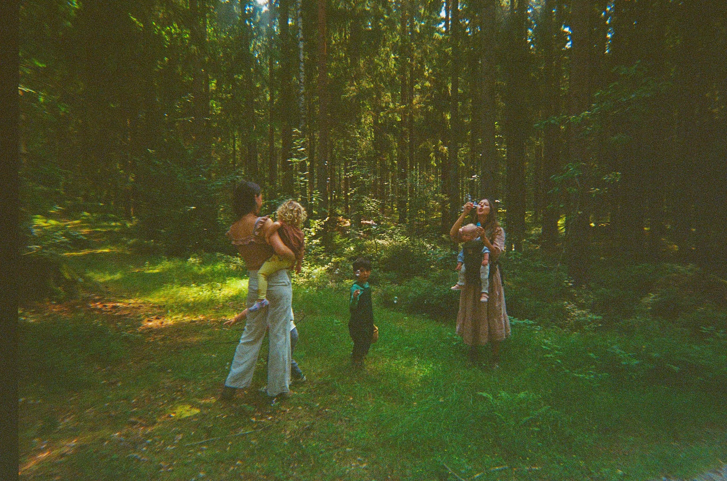 Group of five people, two women and three children, in a forest with tall trees, some of the women holding children, others standing and observing.