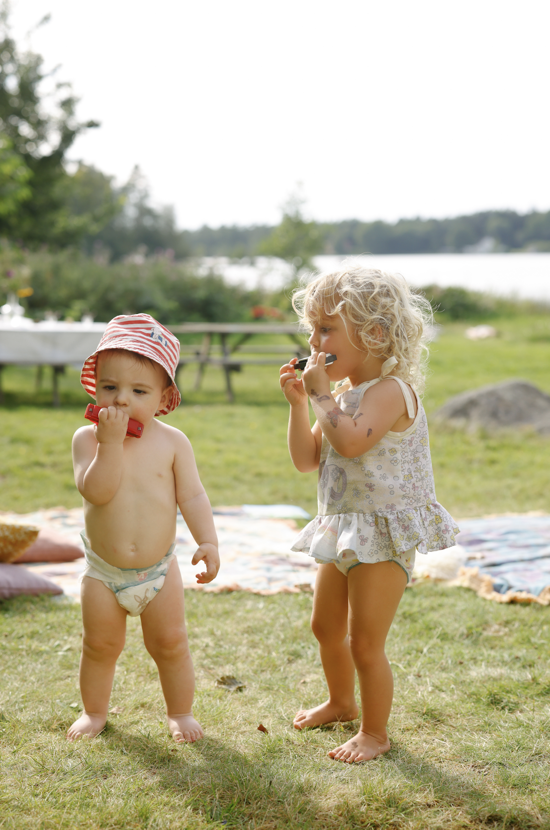 Two young children, a toddler and a girl, playing outdoors on grass near a lake, holding toy harmonicas, with picnic blankets and trees in the background.