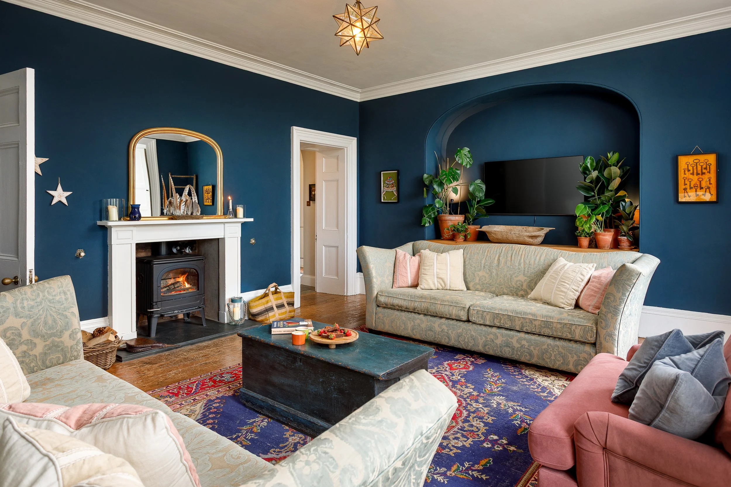 Living room with navy blue walls, a white fireplace with candles on mantel, a mirror above fireplace, a black TV mounted above a beige patterned sofa, a pink armchair, a wooden coffee table, travel books, dishes, a colorful rug, potted plants, framed