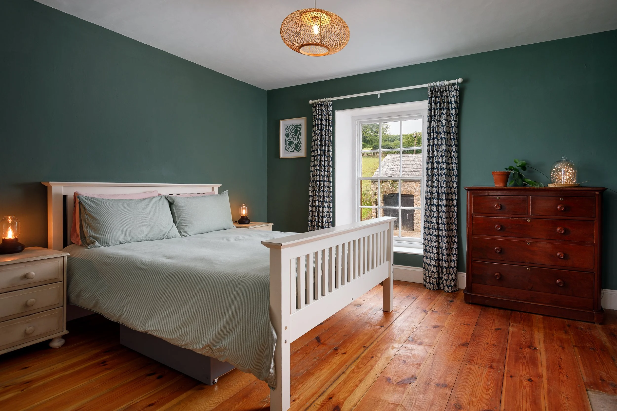 A cozy bedroom with a white wooden bed, striped pillows, nightstands with bedside lamps, a large window with patterned curtains, a framed wall art, and a tall wooden dresser with a potted plant and decorative item, hardwood floors, and a woven ceiling light
