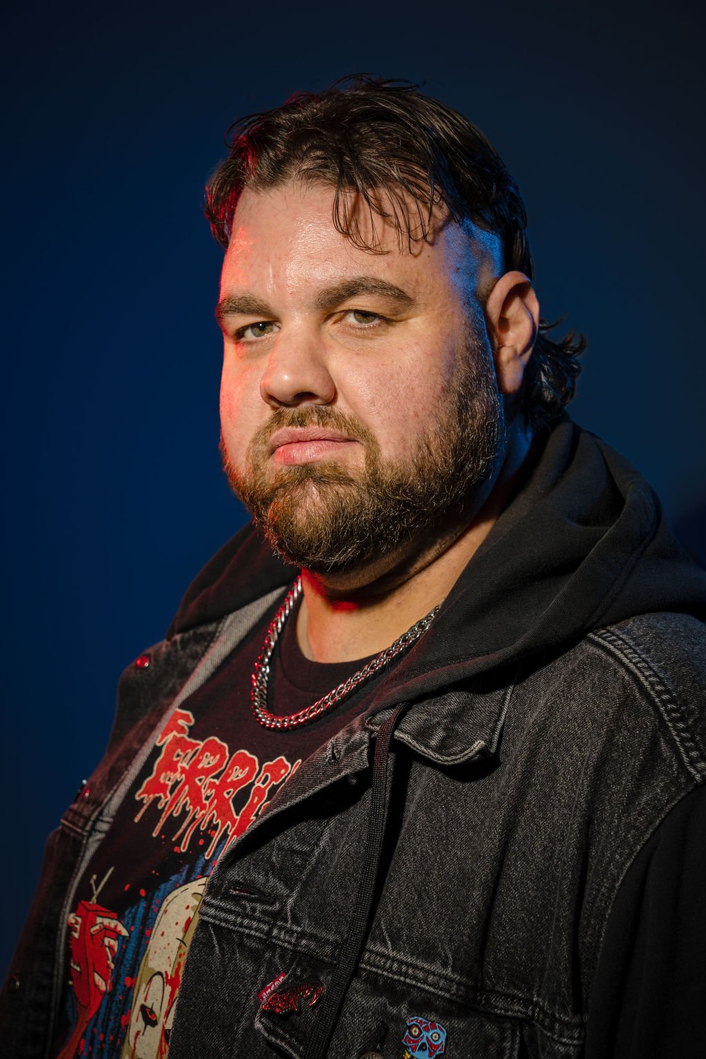 A man with dark hair and a beard wearing a black jacket, a graphic T-shirt, and a silver chain, looking confidently at the camera against a dark blue background.