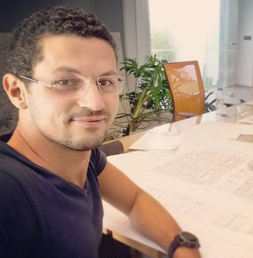 A man with glasses and curly hair sitting at a desk with papers, in a bright office with a large window and a plants.