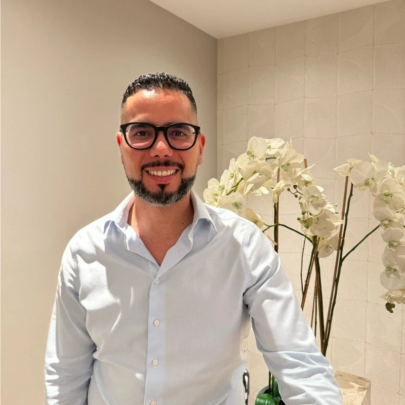 Man with glasses and beard smiling in a white shirt, standing in front of a wall with white orchids.