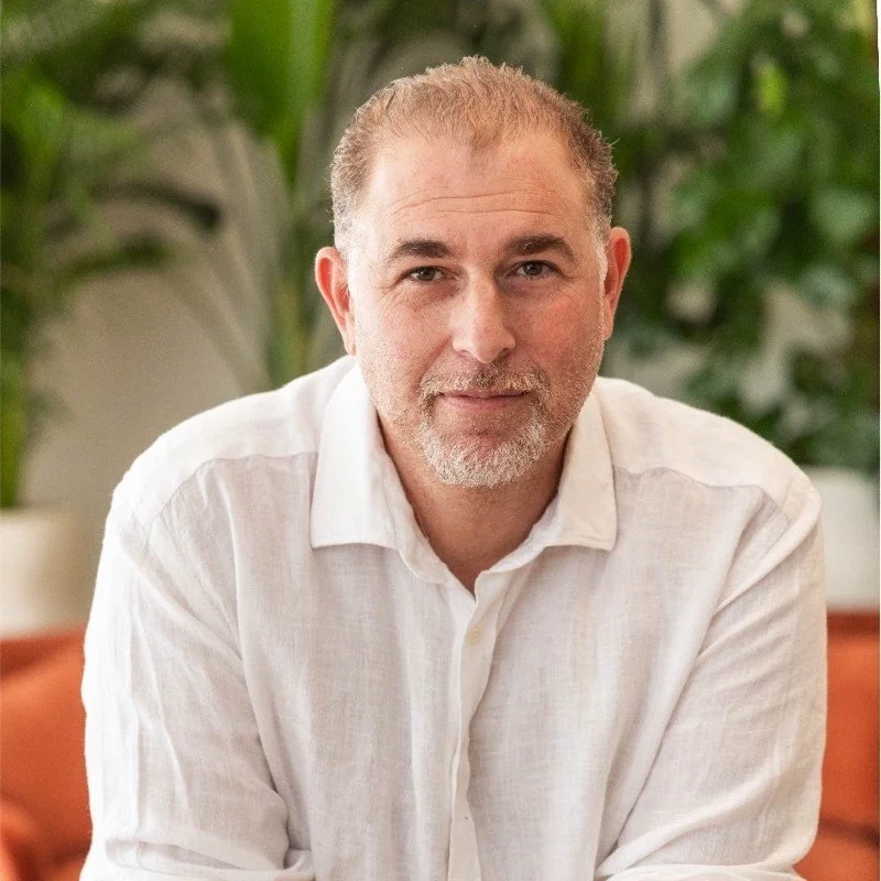 A middle-aged man with short, light brown hair, a beard, and mustache, wearing a white button-up shirt, sitting in a room with green plants and a beige wall in the background.