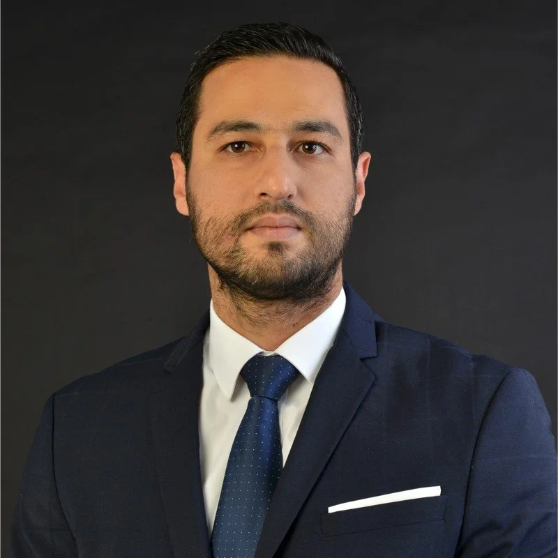 A man with dark hair and beard dressed in a navy suit, white shirt, and navy tie standing against a dark background.