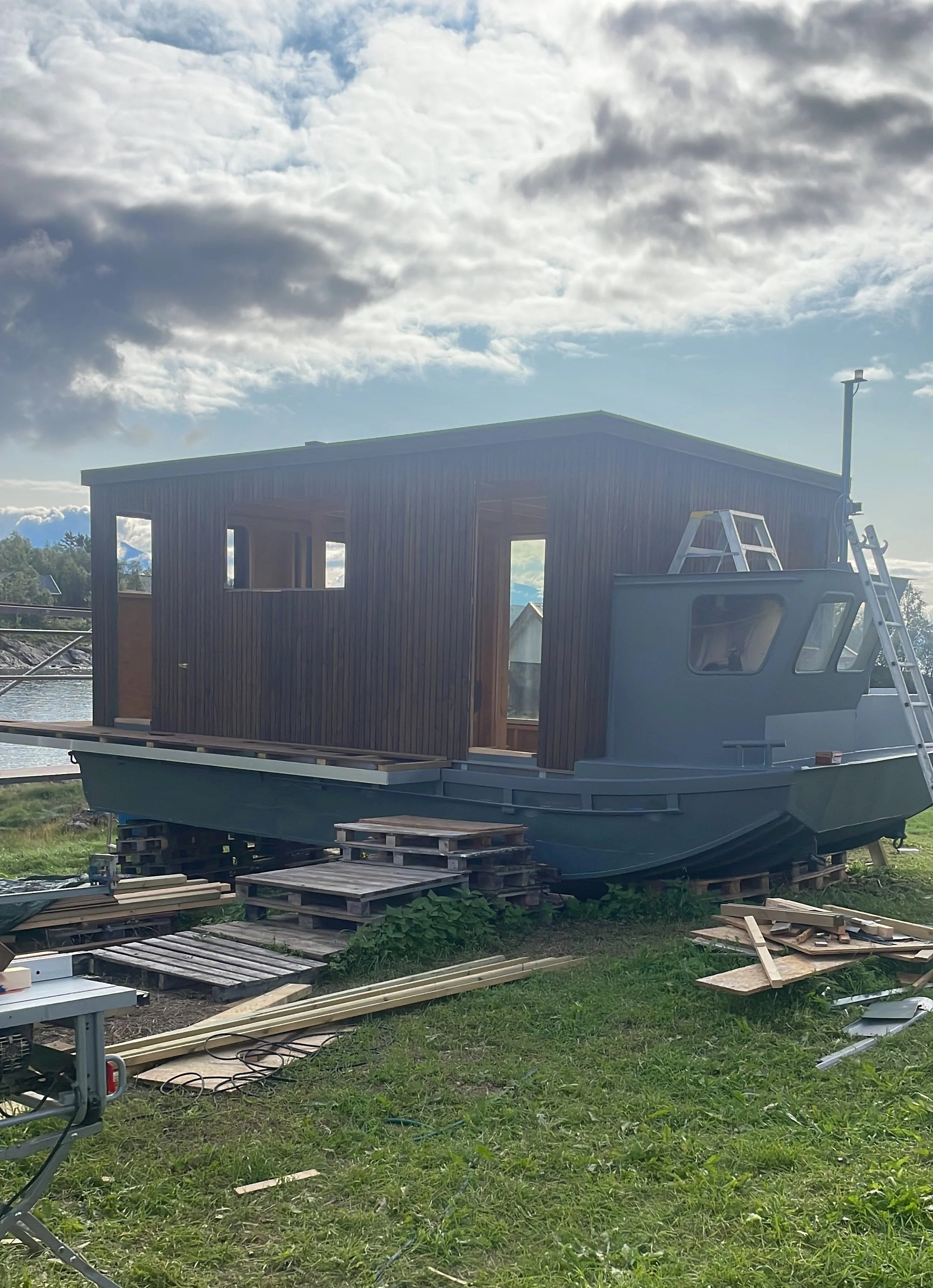 Houseboat under construction on land with wood planks and ladders, grassy area, and cloudy sky.