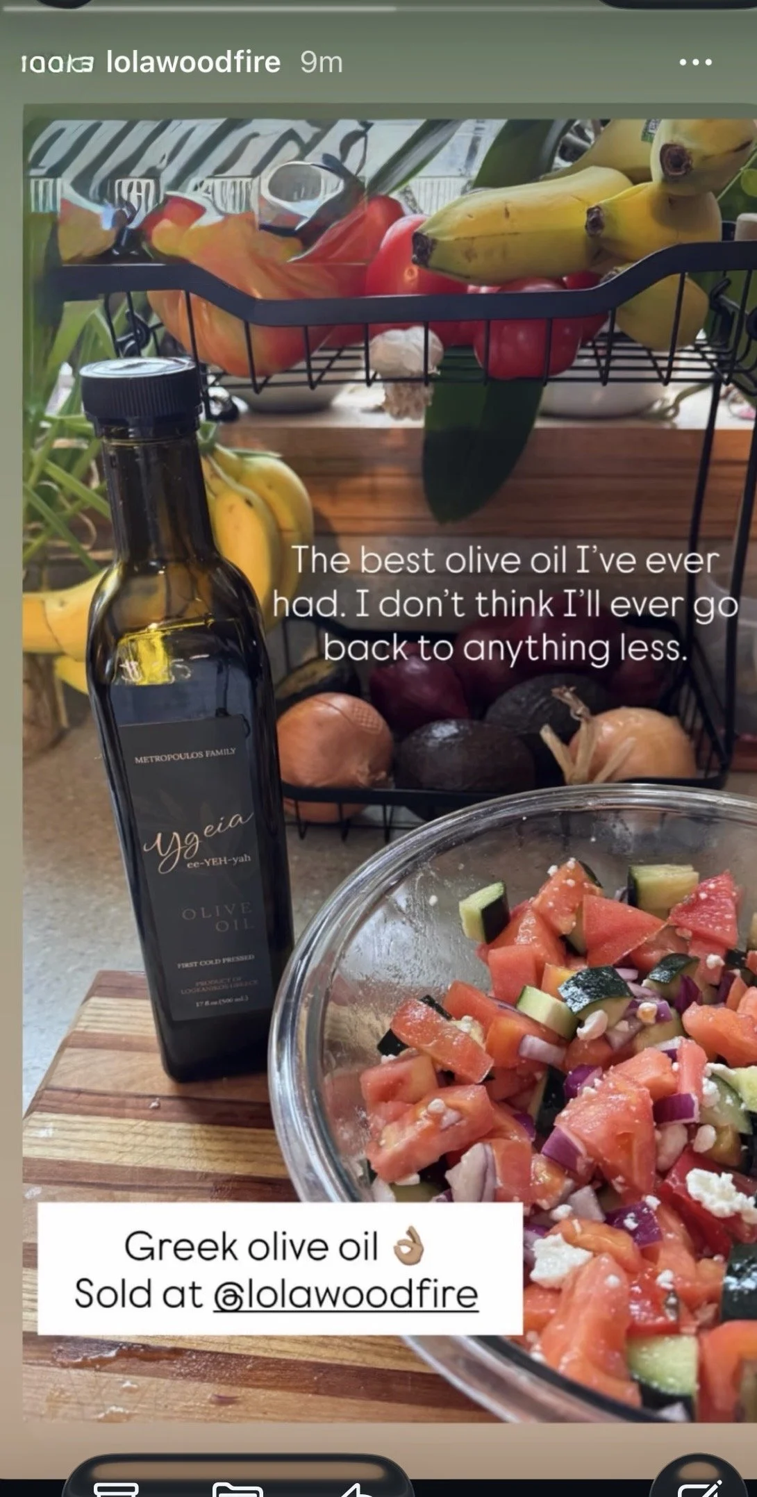A bottle of Greek olive oil, a bowl of chopped salad with tomatoes, cucumbers, onions, and feta cheese, and a basket of bananas, apples, and onions on a kitchen counter.