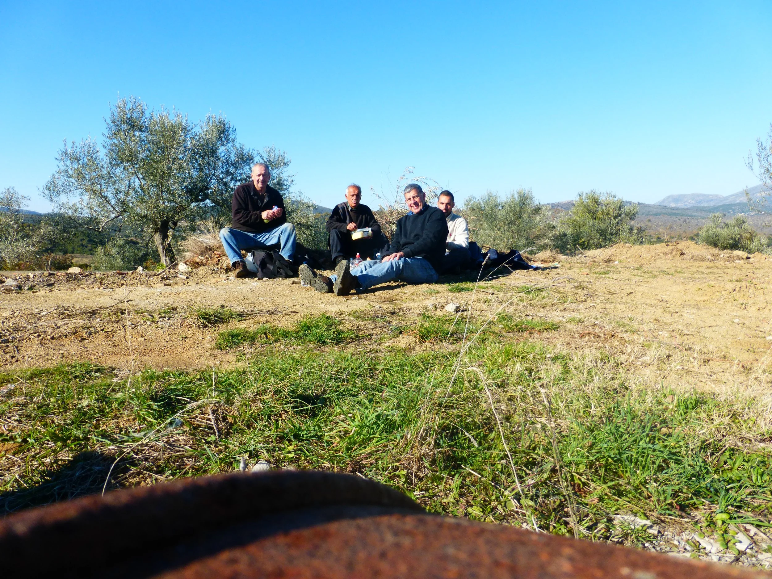 Members of the Metropoulos family taking a break during the olive harvest in their family grove in Logkanikos, Laconia, Greece, surrounded by Athinolia olive trees and the rocky mountain landscape.