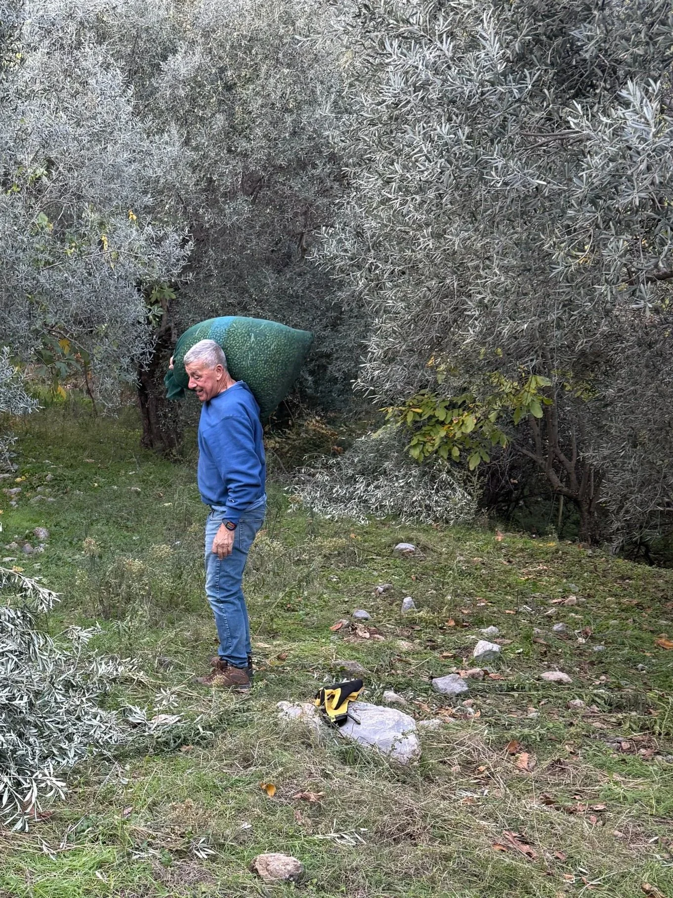 A man with gray hair, wearing a blue jacket, jeans, and brown shoes, standing on a grassy area surrounded by trees, carrying a large green bag on his shoulder, smiling. A yellow and black pair of gloves are on the ground near his feet.