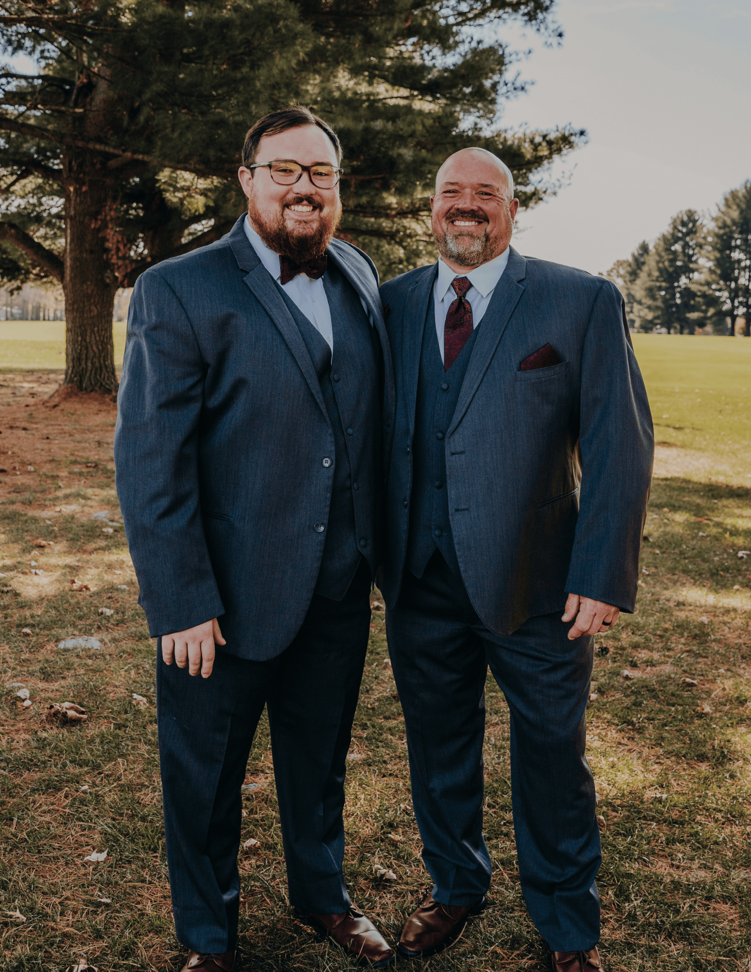 Two men in formal suits standing outdoors on grass with trees in the background.