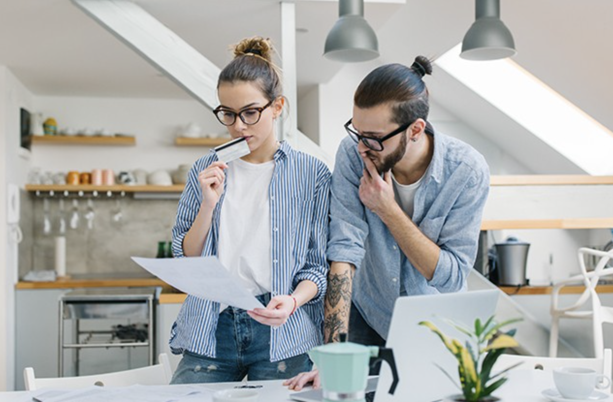 Deux jeunes adultes, un homme et une femme, regardent un document dans une cuisine moderne, la femme tient une barre de chocolat à côté de la bouche, tous deux semblent concentrés sur le document.