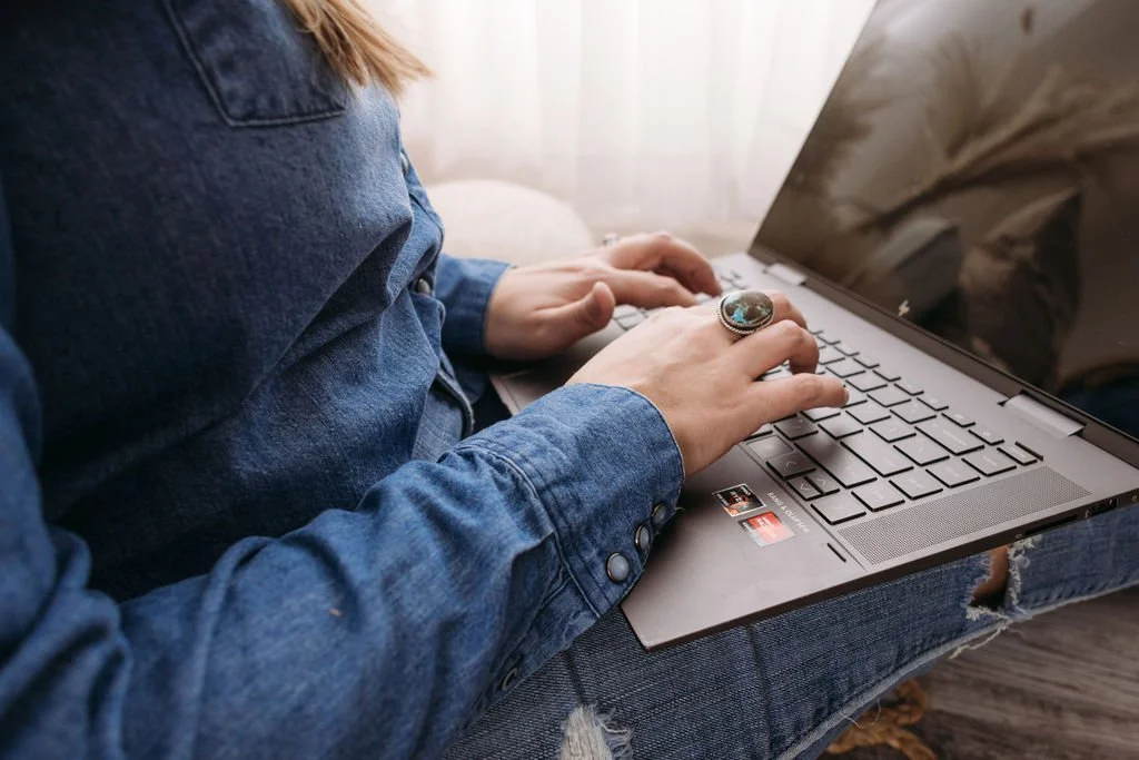 A person wearing a denim jacket and ripped jeans is sitting on a sofa using a laptop, with one hand on the keyboard and a large ring on the finger of the hand on the keyboard.