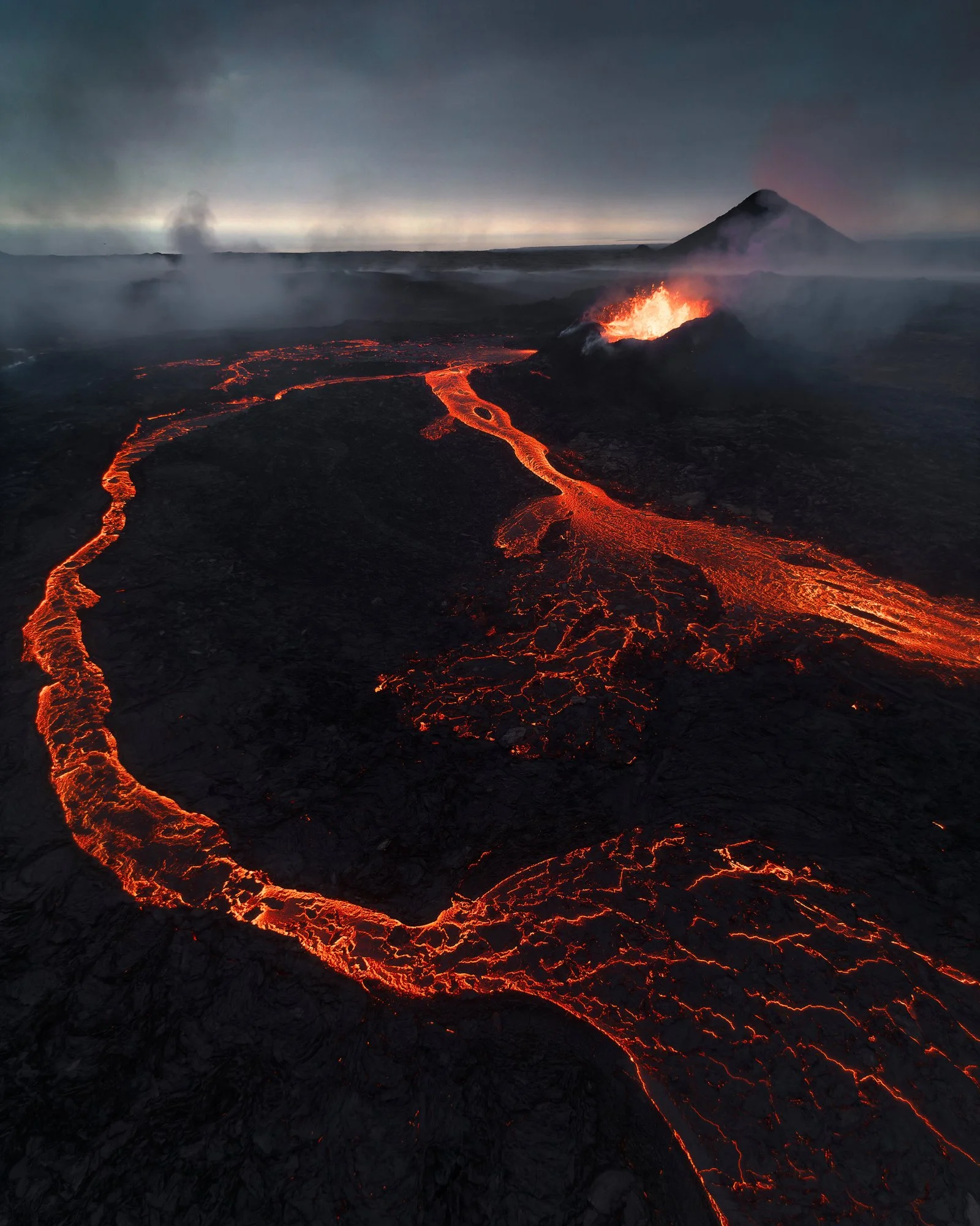 A mesmerizing view of an active volcanic eruption, with molten lava rivers glowing intensely against the dark, rugged terrain. The fiery lava flows carve dramatic paths through the blackened landscape, radiating heat and energy. In the background, th