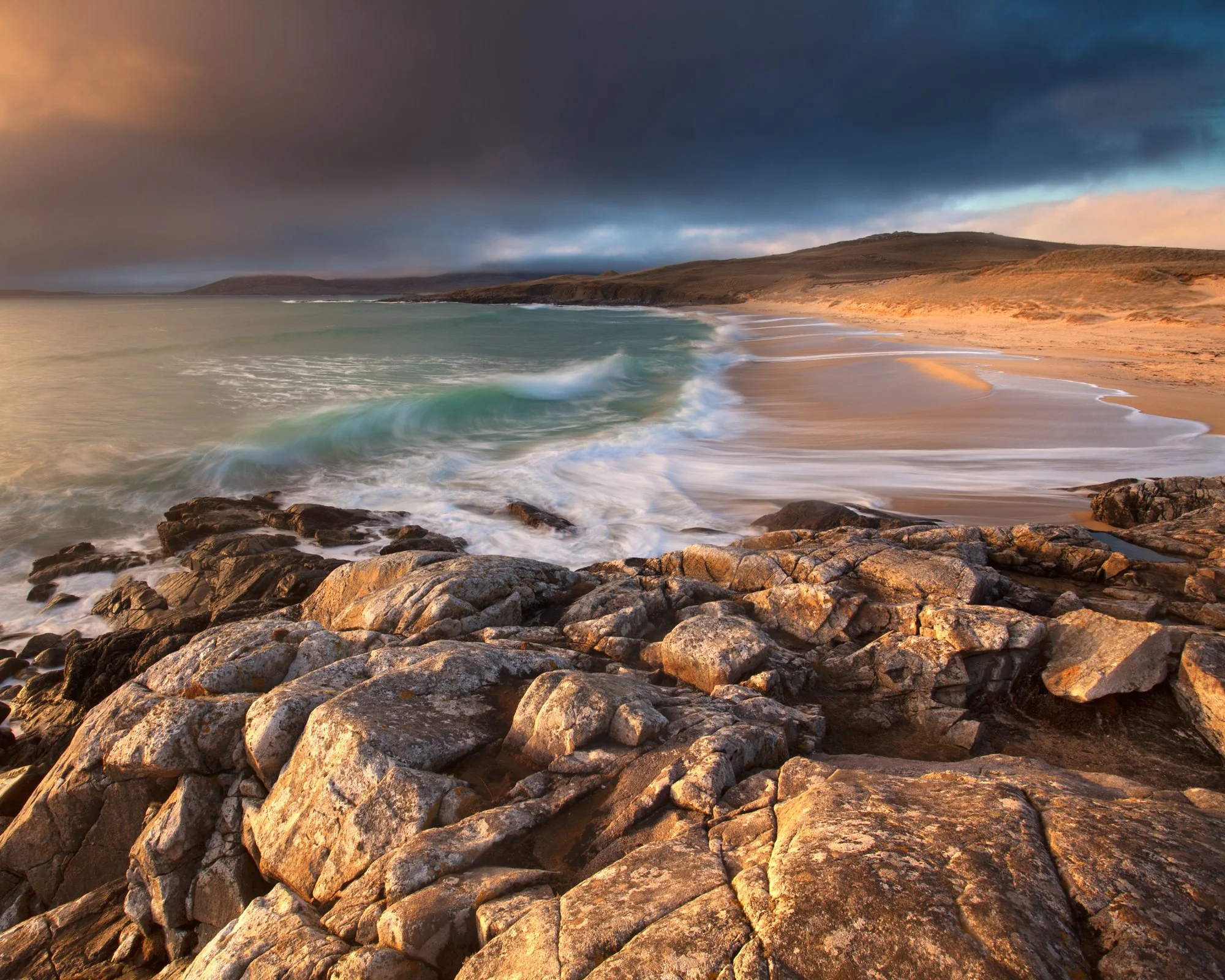 A stormy yet vibrant scene unfolds at Nisabost Beach (Traigh Iar) on the Isle of Harris, with the golden hues of the setting sun illuminating the rugged foreground rocks. The turquoise waves roll onto the shore in gentle arcs, contrasting beautifully
