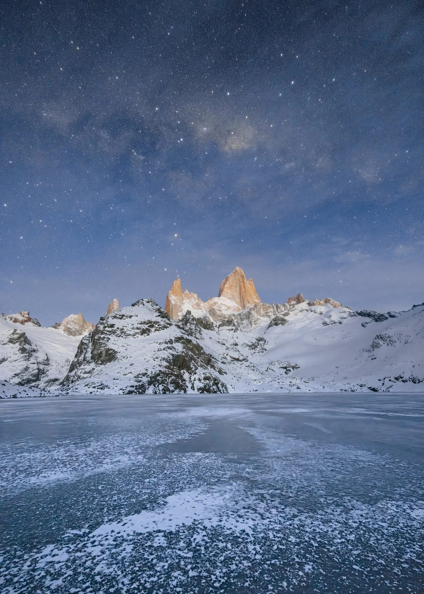 Beneath a sky filled with countless stars, the iconic peaks of Mount Fitz Roy rise majestically above the frozen surface of Lago de los Tres in Patagonia. The snow-draped mountains catch the faint glow of starlight, creating a stunning contrast again