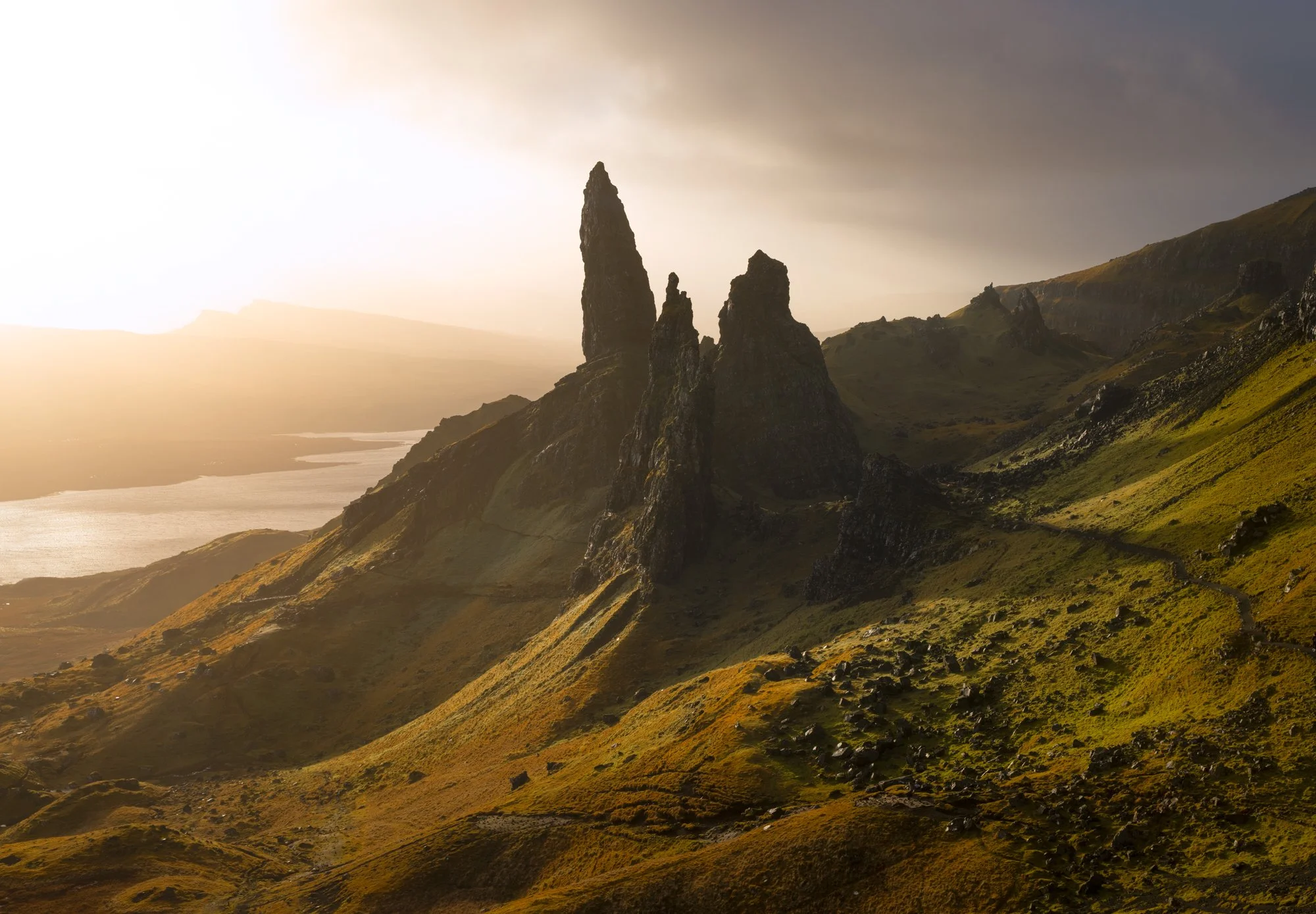 The Old Man of Storr rises dramatically from the rugged landscape of the Isle of Skye, illuminated by the warm, golden glow of side light at sunrise. Shadows stretch across the vibrant green hills, adding depth and contrast to the iconic rock formati