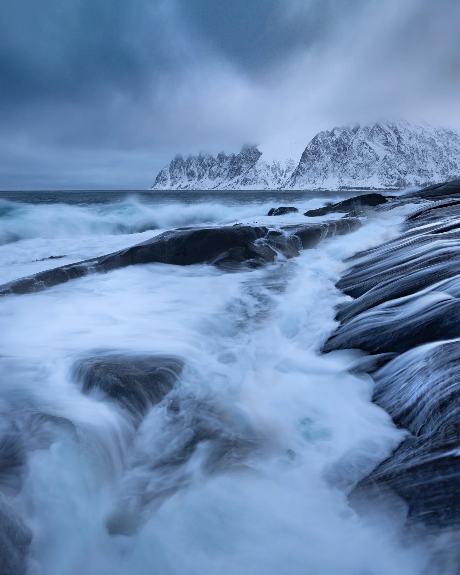 Angry waves crash against the rugged coastline of Tungeneset on Senja Island, creating dynamic, sweeping patterns of foam that draw the eye toward the distant, snow-covered mountains. Captured using combined exposures, the photograph highlights the d