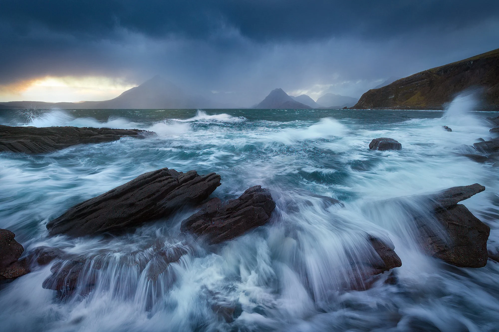Elgol’s dramatic coastline is on full display as waves crash powerfully against dark, jagged rocks under a moody sky. The turbulent sea, captured with perfect motion blur, contrasts beautifully with the brooding mountains in the distance. Over twenty
