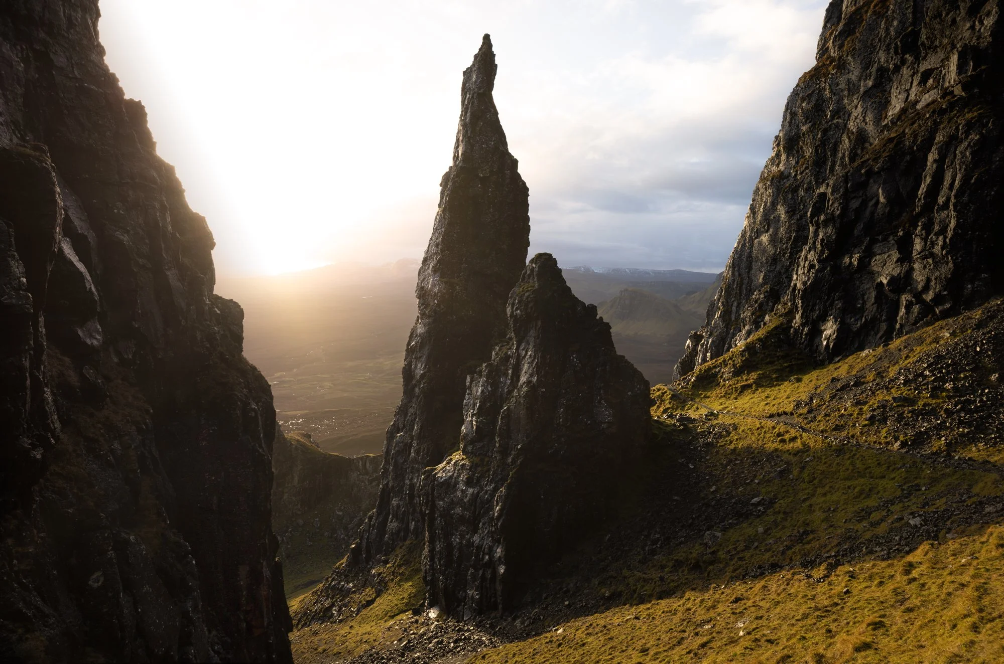 The jagged rock formation known as The Needle stands tall, bathed in the golden glow of low sunlight breaking through the clouds. The contrasting dark shadows of the surrounding cliffs add depth and drama to the scene. Captured at the Quiraing on the