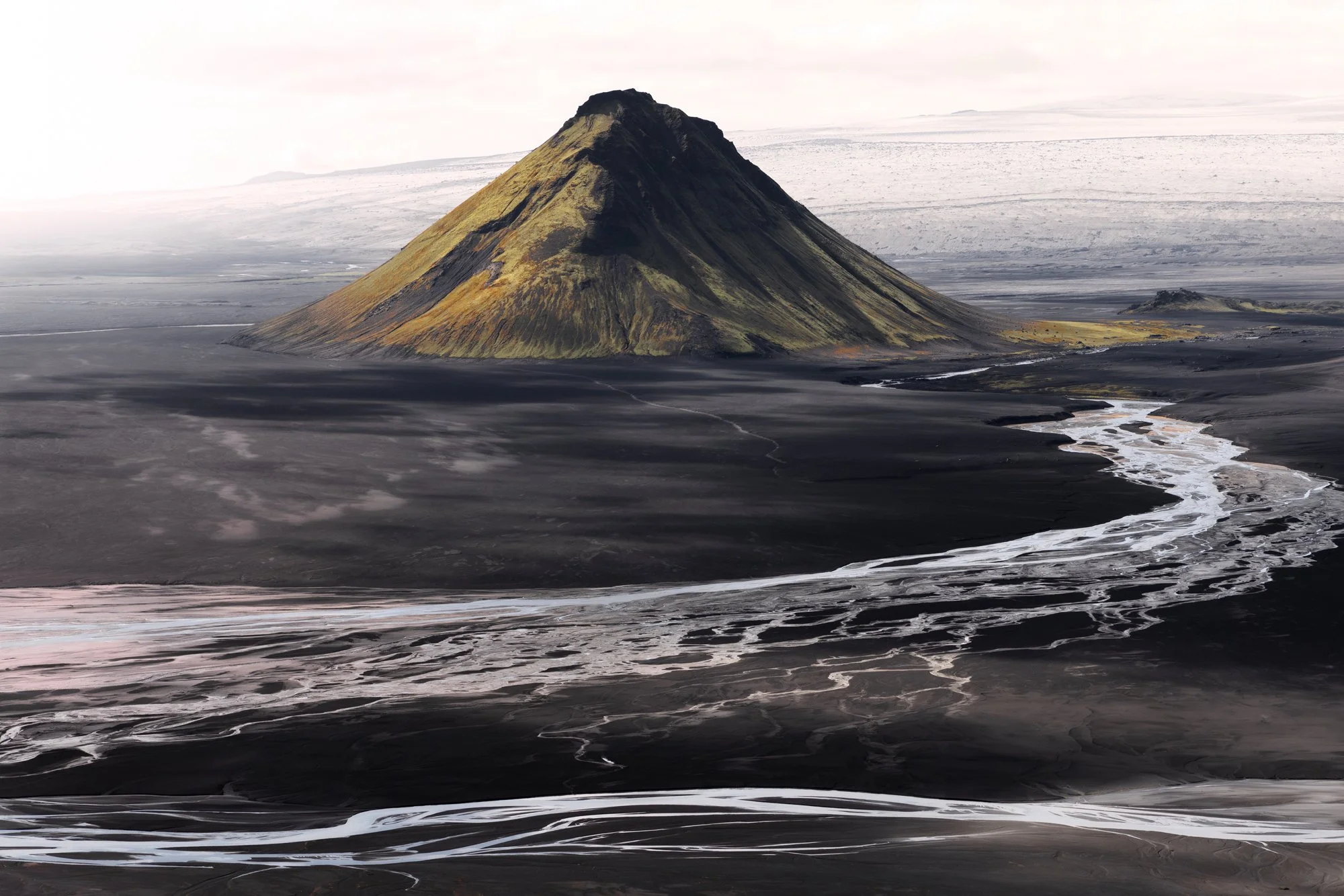 An isolated volcanic peak stands prominently amidst a vast expanse of black sand flats, with streaks of glacial meltwater weaving intricate patterns across the dark desert floor. Mælifell’s vivid green moss clings to its slopes, providing a striking 