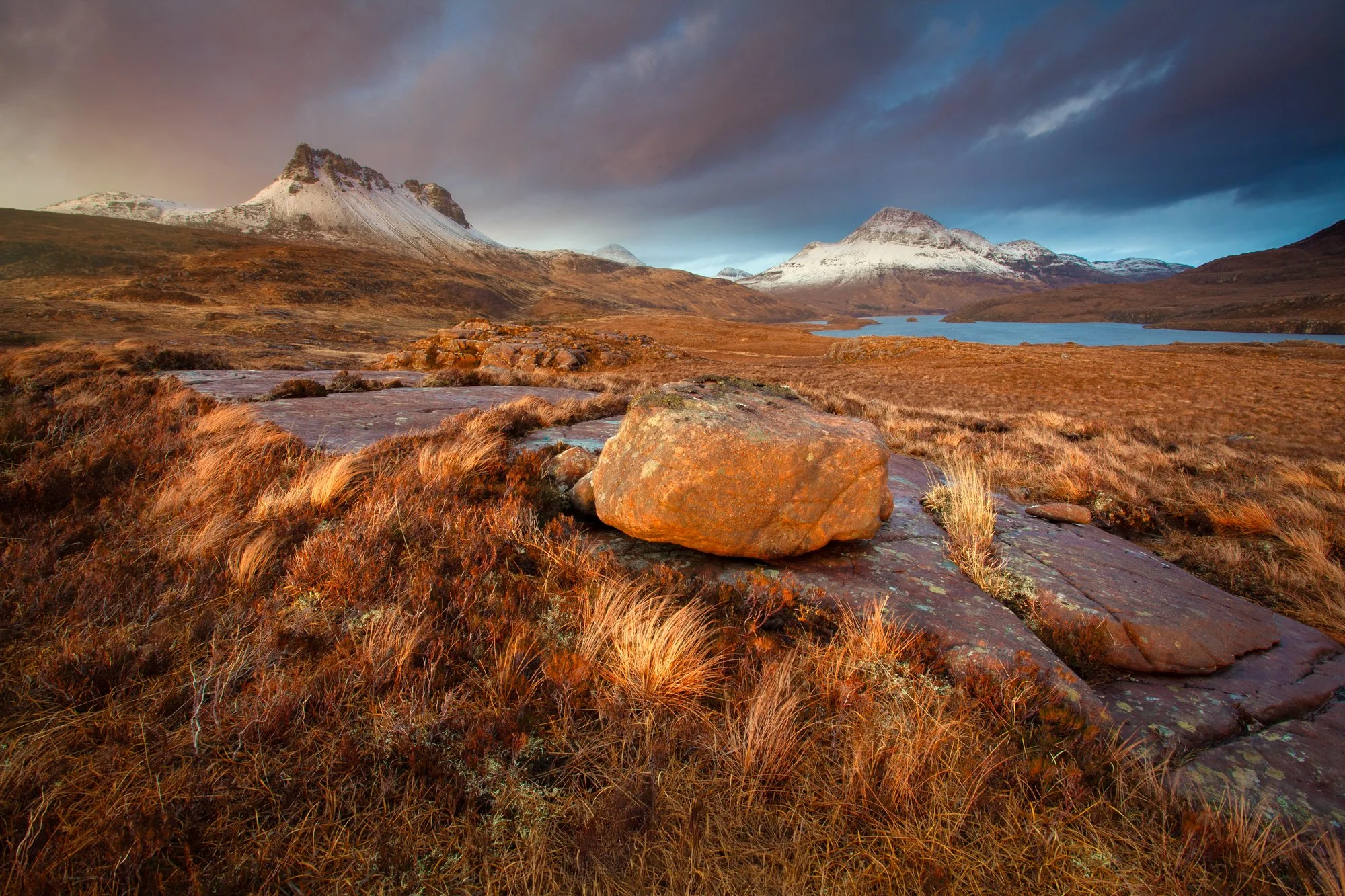 A stunning sunset illuminates the rugged Scottish Highlands with the twin peaks of Stac Polliadh and Cùl Beag standing out majestically. The warm hues of golden light enhance the textures of the rocky landscape and wild grass in the foreground. Snow-