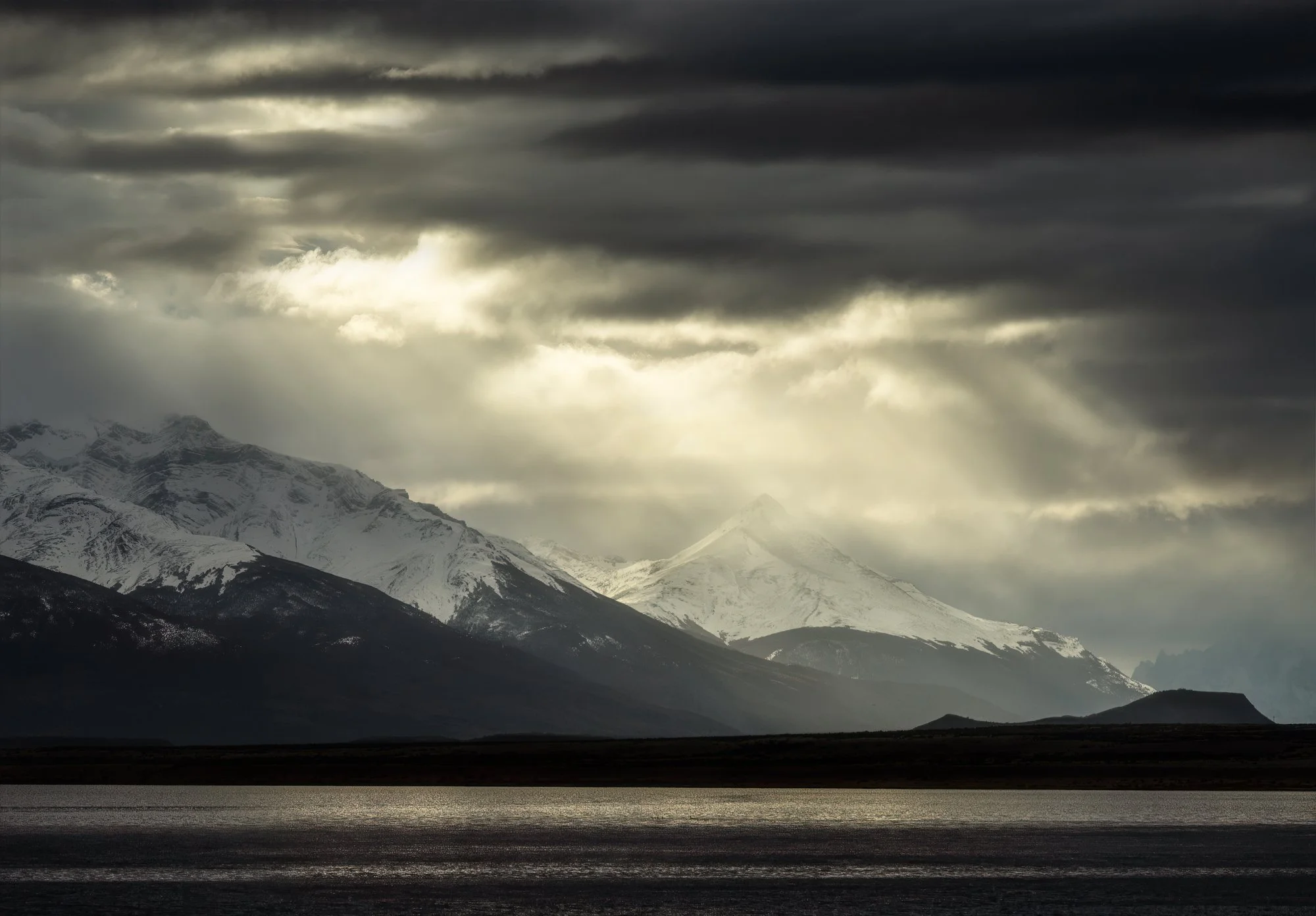 
A majestic scene of the Patagonian mountains captured during a break in the heavy clouds. Beams of light pour dramatically over the snow-dusted peaks, illuminating patches of the rugged terrain below. The foreground features a tranquil body of water