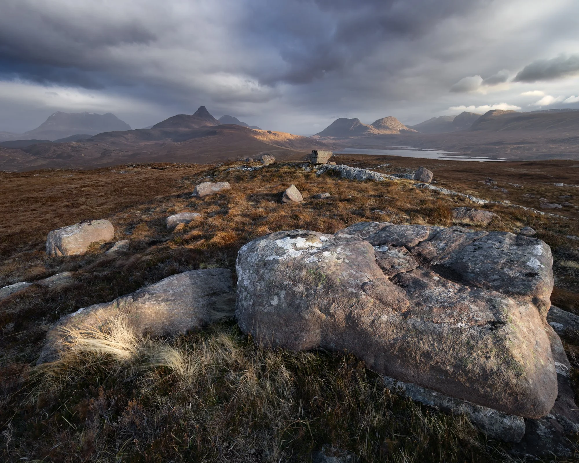 A breathtaking view of Assynt’s wild landscape, with sculpted peaks like Stac Pollaidh and Quinag rising in the distance under a dramatic sky. The golden light softly illuminates the foreground rocks and the expansive heathland, creating a striking c