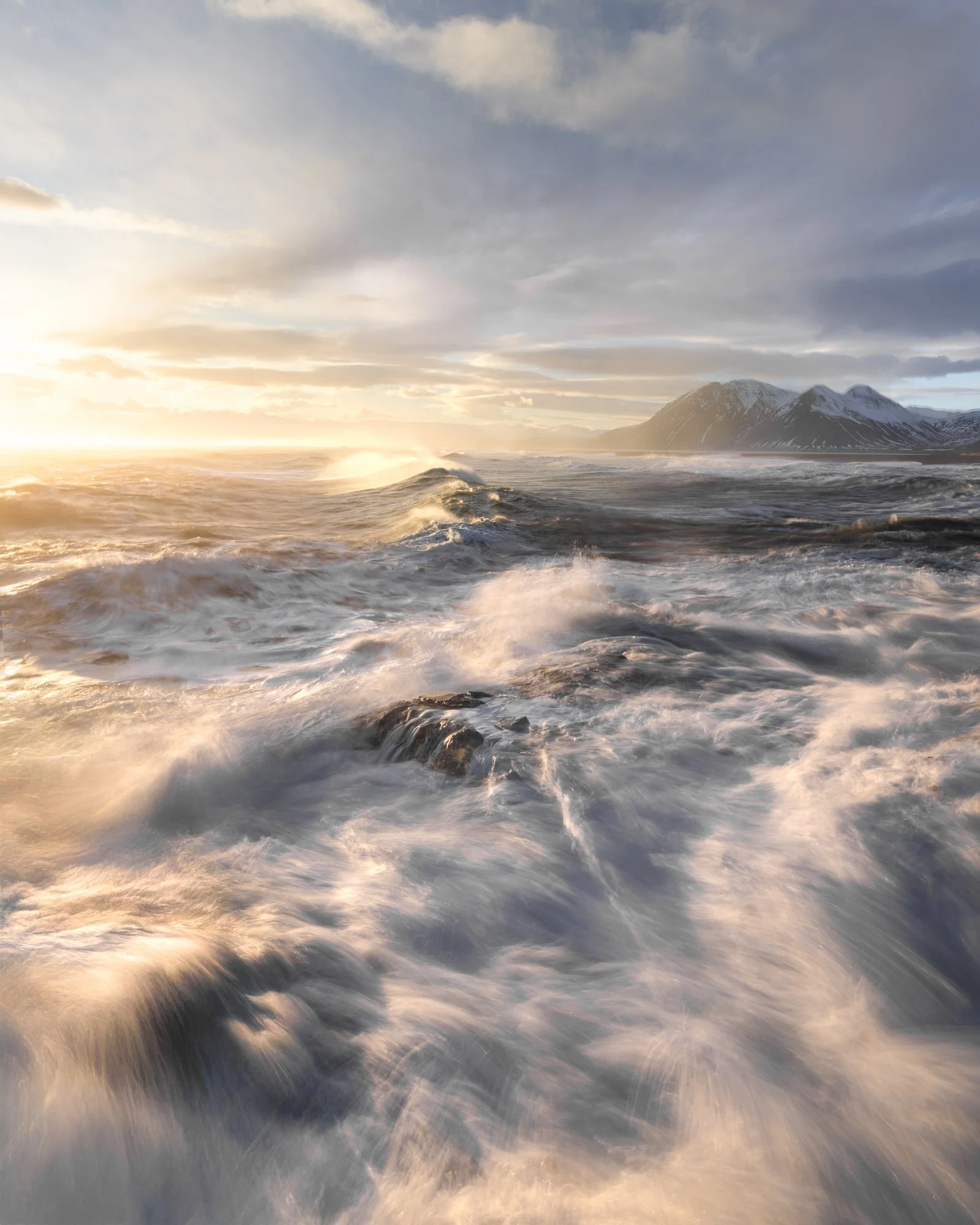 The fierce waves of Iceland’s coastline crash against the rugged shore under a dramatic sky, illuminated by soft, golden light. The wind howls across the sea, whipping up spray and foam, creating a dynamic scene that perfectly encapsulates the wild, 