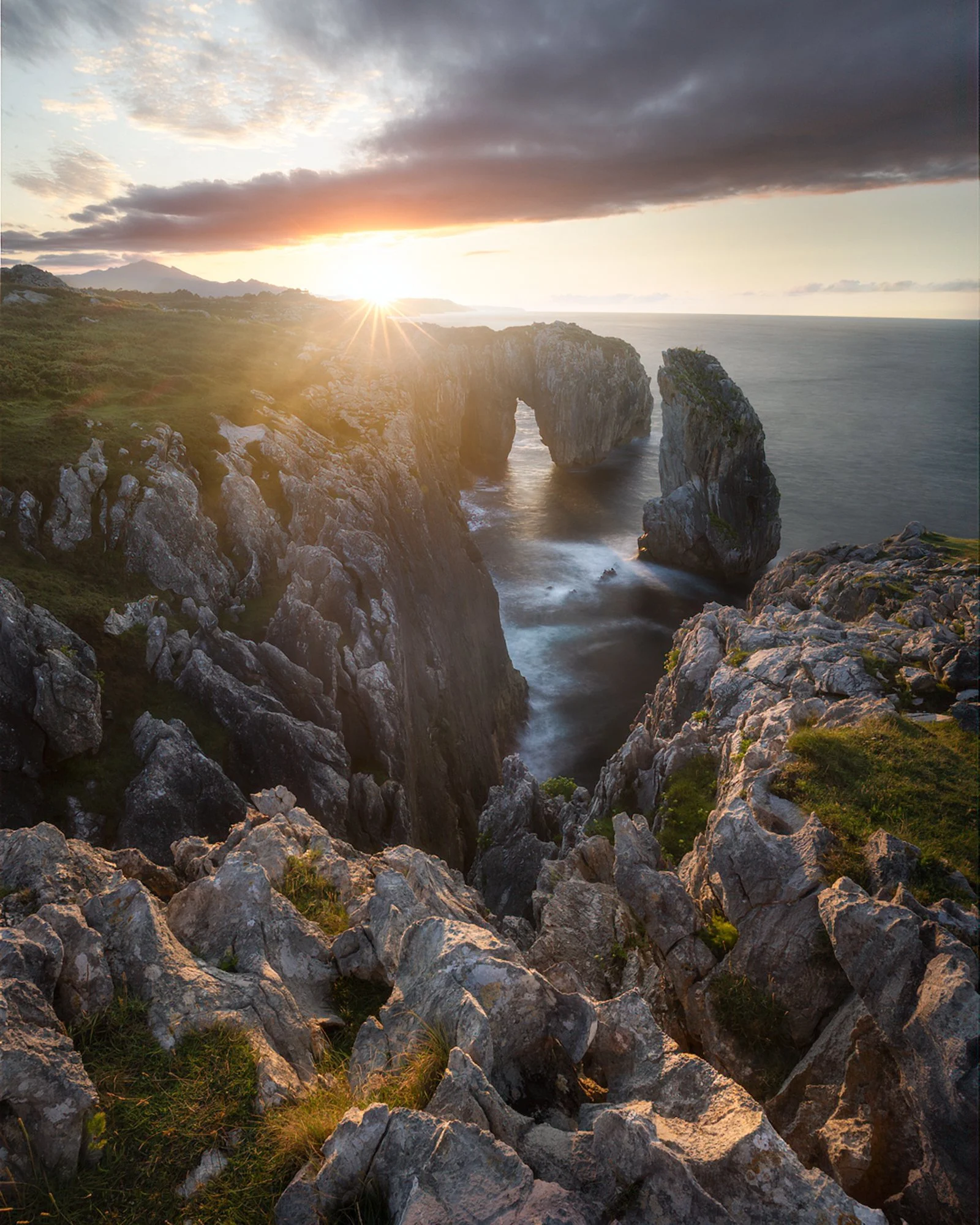 
Golden sunlight pierces through the rugged cliffs of the Spanish coastline, casting a warm glow across the rocky landscape. The natural arch at the center of the frame stands tall against the shimmering sea, while soft, rolling clouds add depth and 