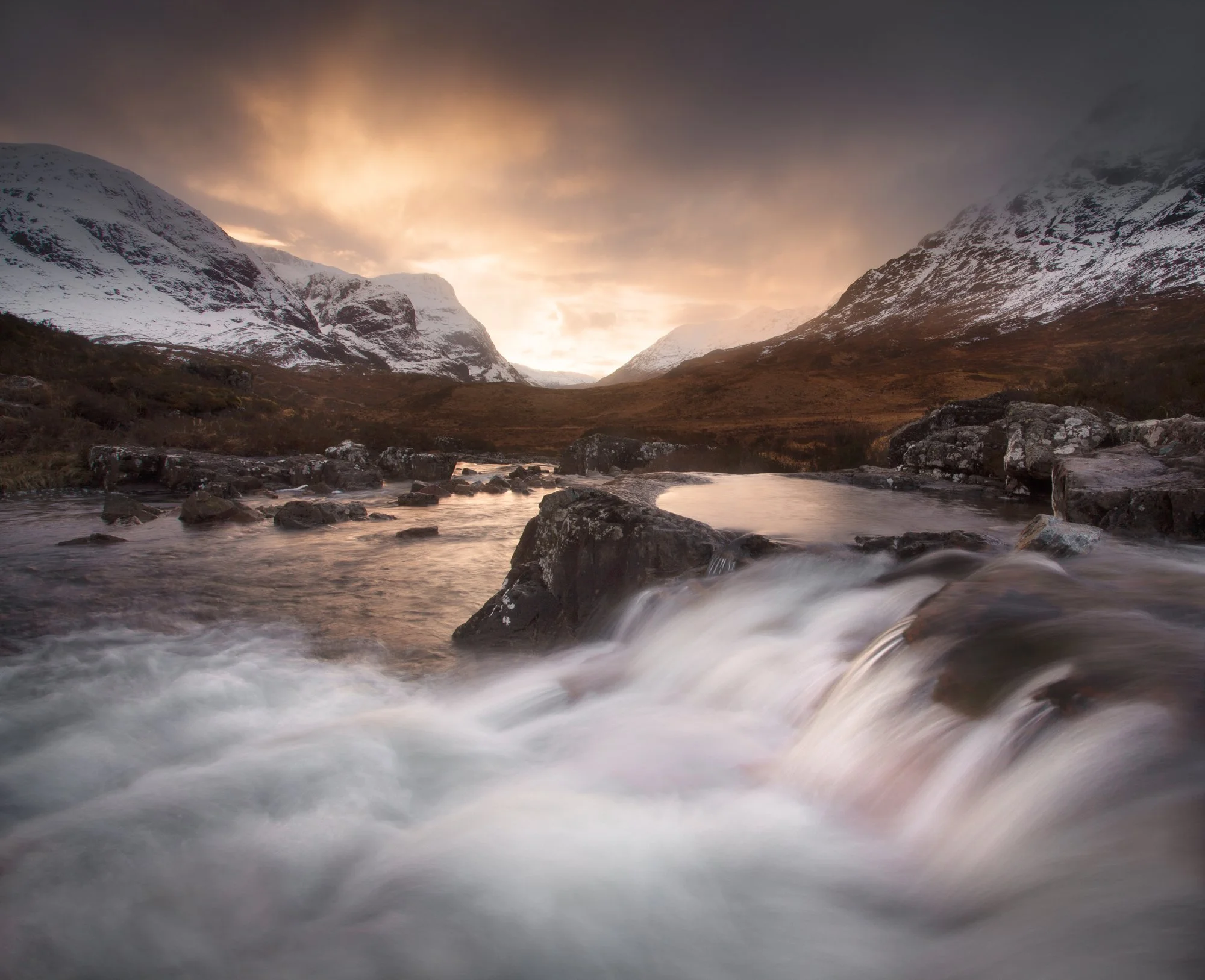 A serene winter landscape in Glencoe captures the golden hues of a sunset gently illuminating the snowy peaks. In the foreground, a rushing waterfall flows gracefully over weathered rocks, its curves mirroring the soft, flowing form of the clouds abo
