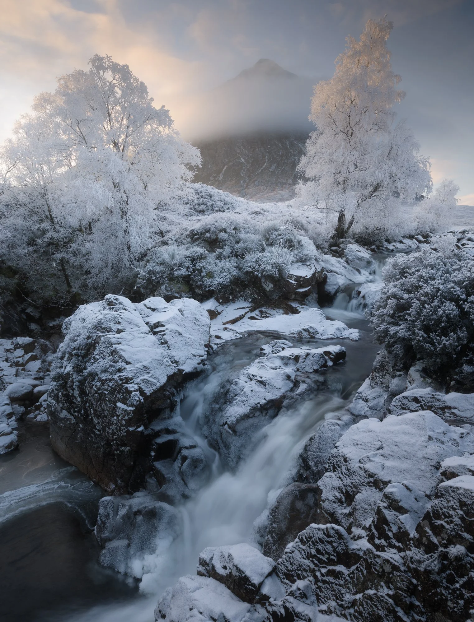 A serene winter landscape at Rannoch Moor, featuring a snow-covered riverbank where the river Coupall flows gently through frozen terrain. In the background, the peak of Buachaille Etive Mòr rises majestically, partially obscured by soft mist. Frost-