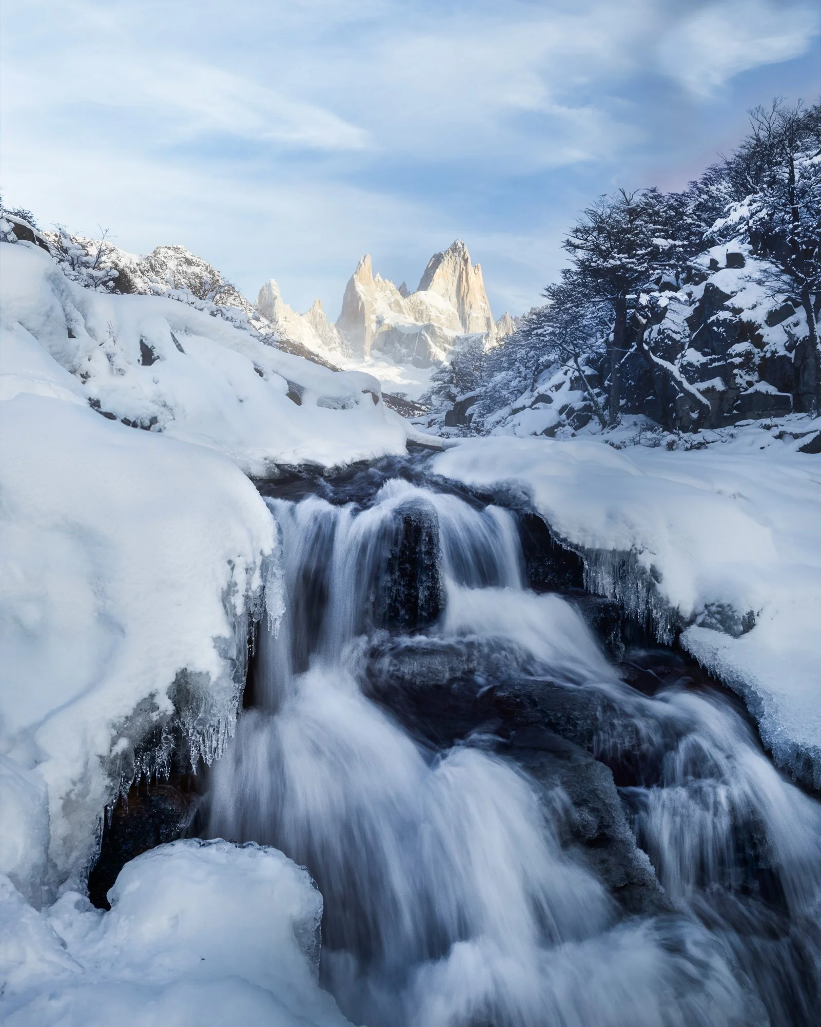 A stunning winter scene captures a frozen waterfall surrounded by deep snow and frost-covered rocks. The cascading water contrasts against the icy landscape, while in the distance, the jagged peaks of Fitz Roy rise majestically under a clear blue sky