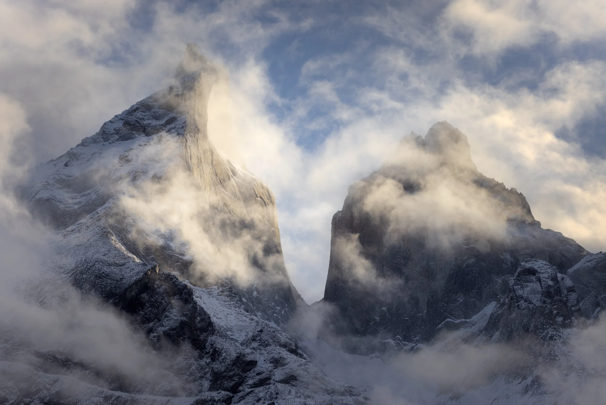 The majestic peaks of the Cordillera Paine emerge dramatically through swirling mist, creating an ethereal atmosphere in Torres del Paine National Park, Chile. The rugged textures of the mountains are highlighted by soft light breaking through the cl