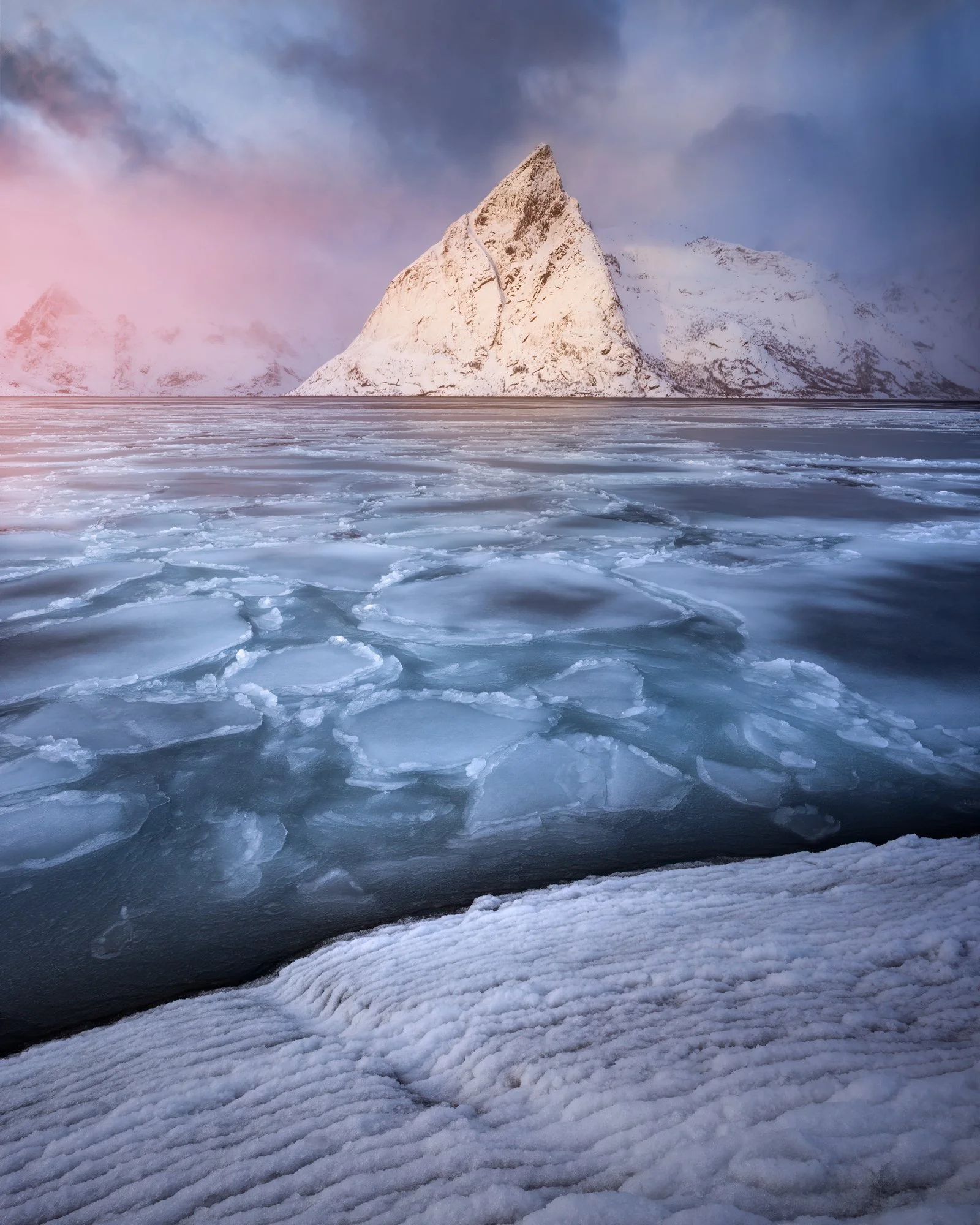 A stunning winter scene in Reine, Norway, with Mount Olstinden standing prominently in the background, framed by delicate layers of sea ice and snow. The soft pink hues of low sunlight gently illuminate the rugged mountain and frozen landscape, creat