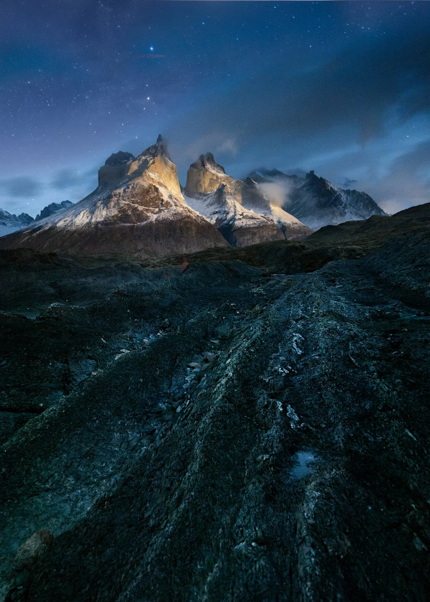 Under a deep night sky filled with stars, the jagged peaks of Torres del Paine are illuminated by an otherworldly moonrise that casts a golden glow resembling sunrise. The foreground, with its rugged textures and dark tones, leads the viewer’s eye to