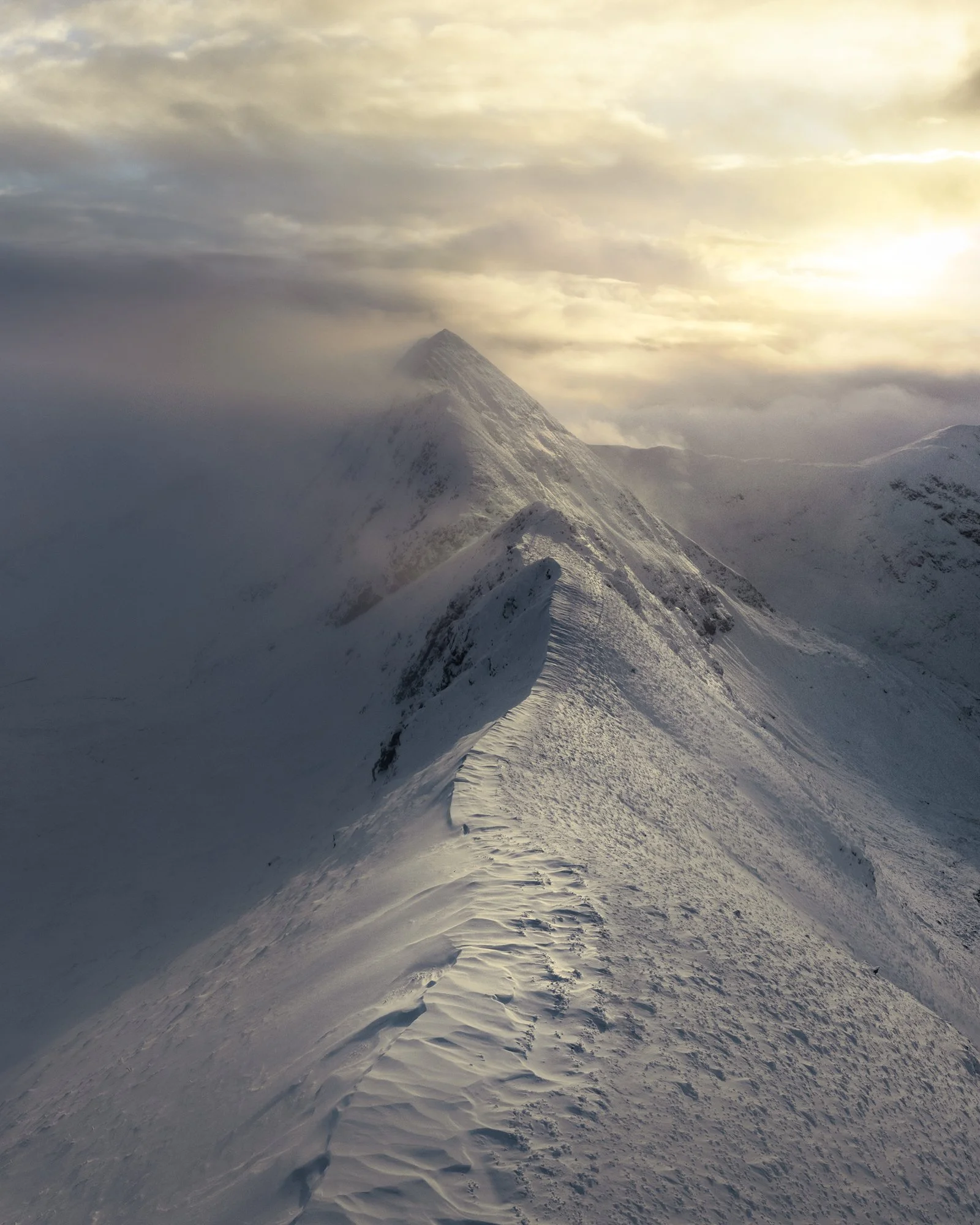 A pristine ridge of snow snakes toward a distant summit, illuminated by soft, golden light breaking through low-hanging clouds. The rugged terrain of Sgùrr a’Mhaim and The Devil’s Ridge creates an awe-inspiring scene, where untouched snowdrifts and s