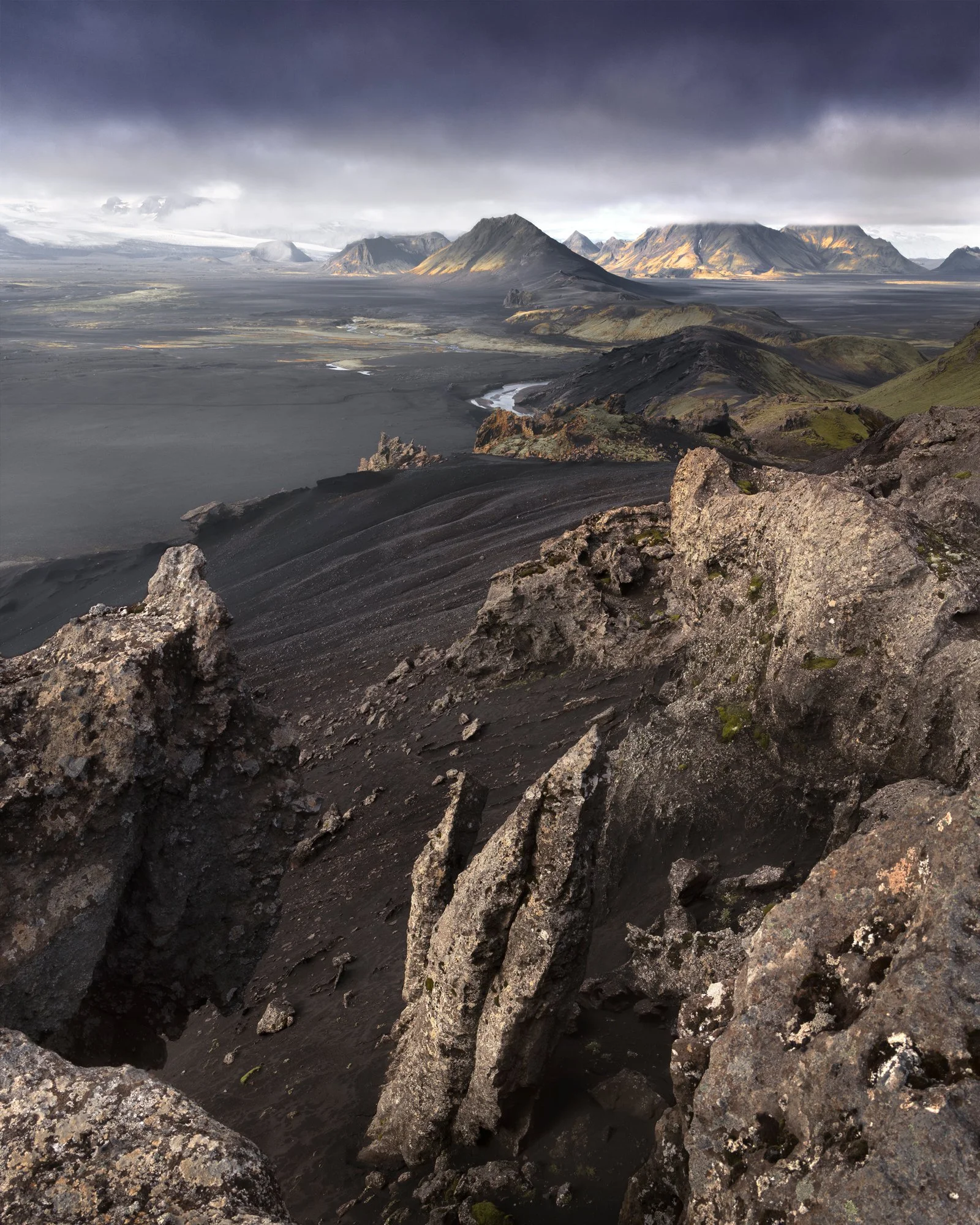 A dramatic landscape unfolds, capturing the start of a six-day trek through the Icelandic Highlands. The foreground features rugged volcanic rock formations, leading the eye towards a vast expanse of black sand flats and river channels snaking throug