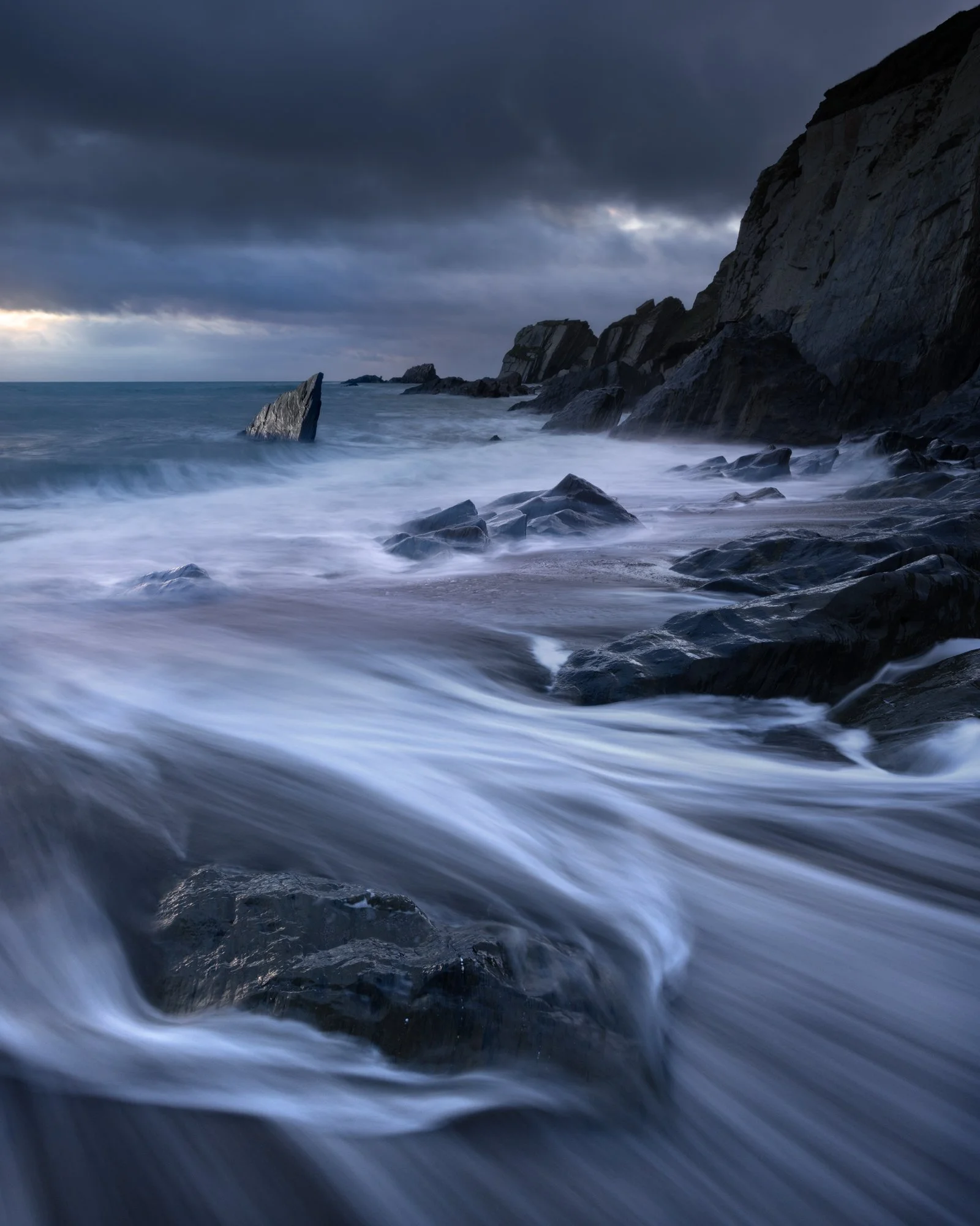 Foamy waves rush over the dark rocks of the South Hams coast, creating elegant leading lines under a moody, overcast sky. The monochrome tones in this late autumn scene emphasize the texture and drama of the rugged shoreline. Captured using multiple 